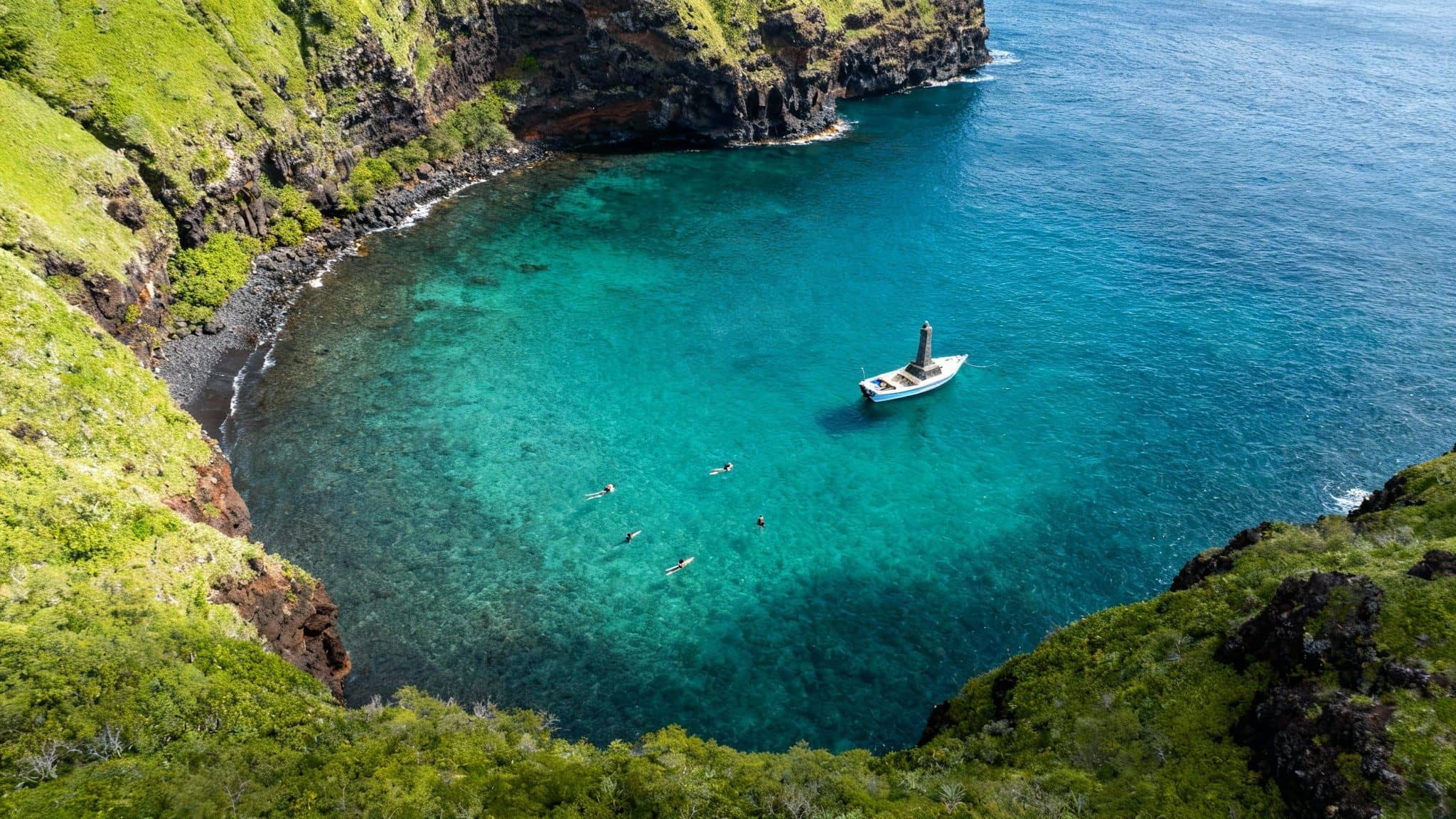Aerial view of a vibrant blue bay with green cliffs, a boat, and snorkelers in clear water.