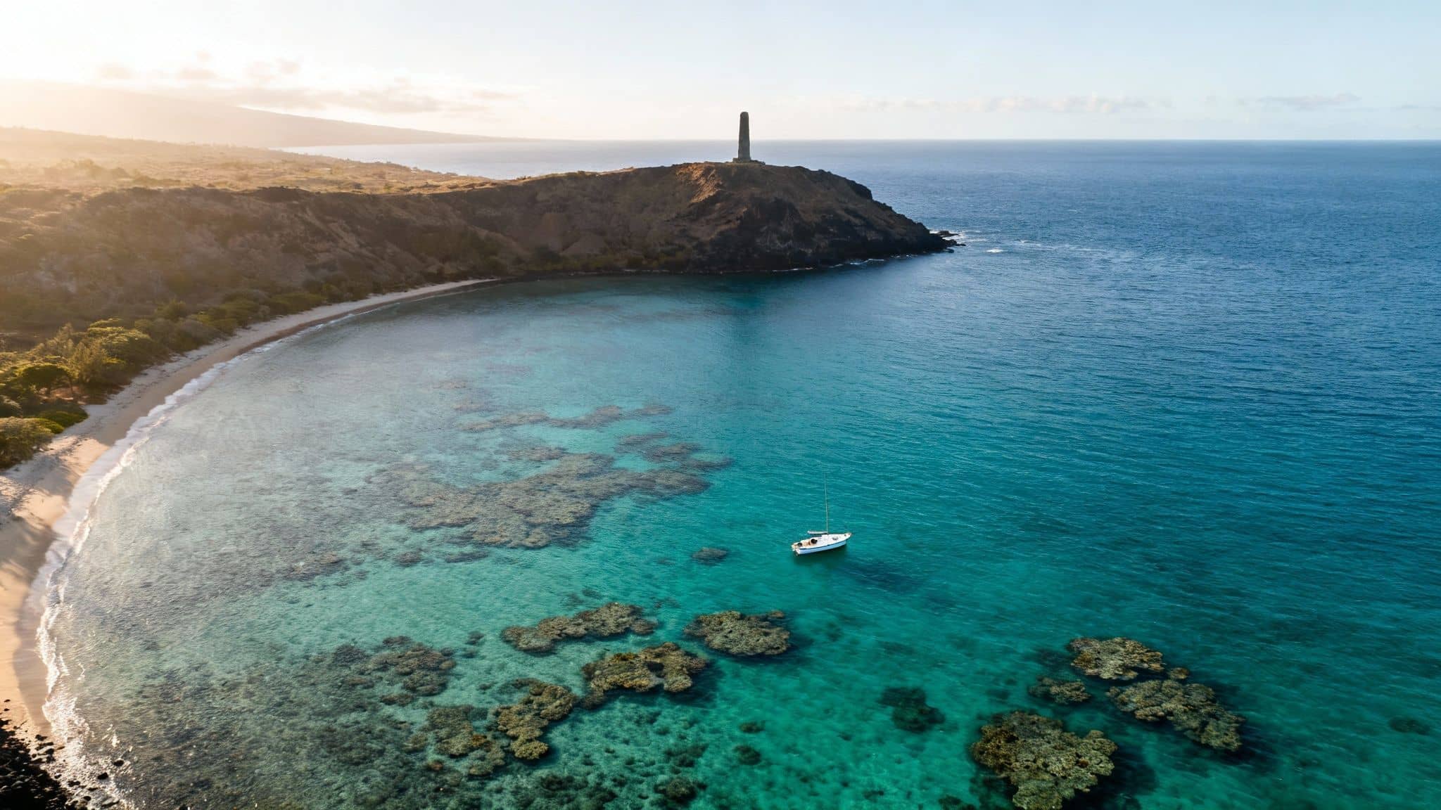 Aerial view of a tropical bay with clear turquoise water, coral reefs, a sandy beach, and a sailboat.