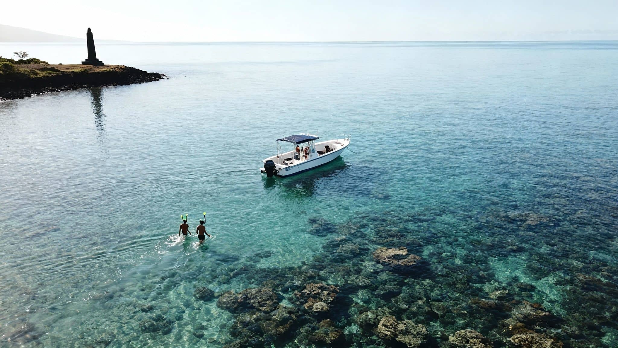 Two people snorkeling in clear turquoise water near a boat, a lighthouse, and underwater coral.