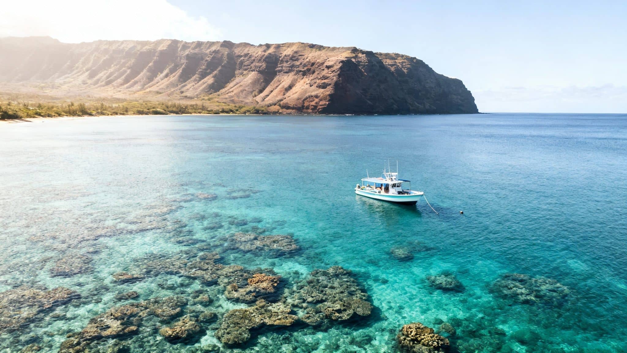 Aerial view of a boat anchored in clear turquoise water near a coral reef and a large mountain.