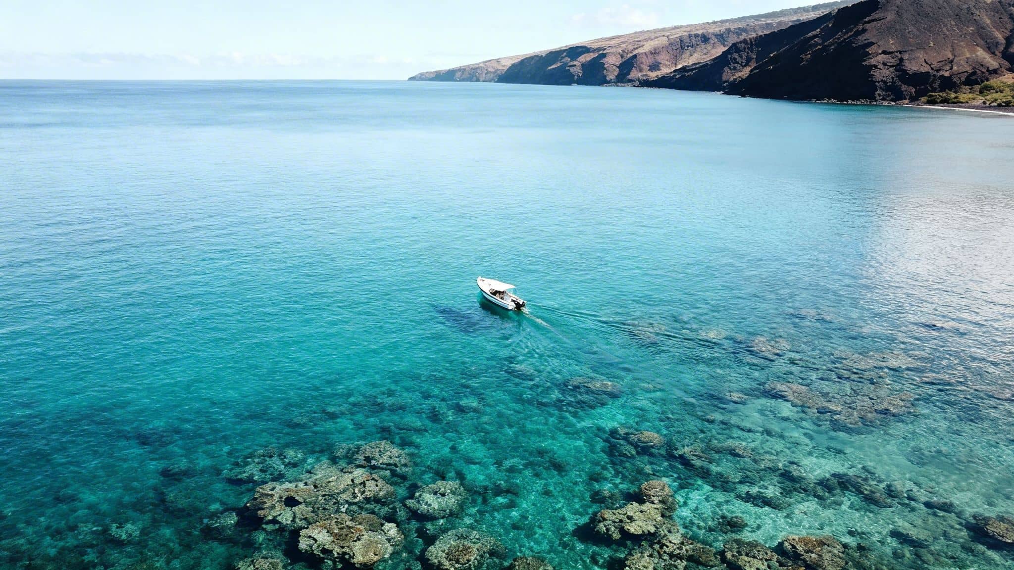 Aerial view of a white boat moving over clear turquoise ocean water with visible coral reefs near a rocky coastline.