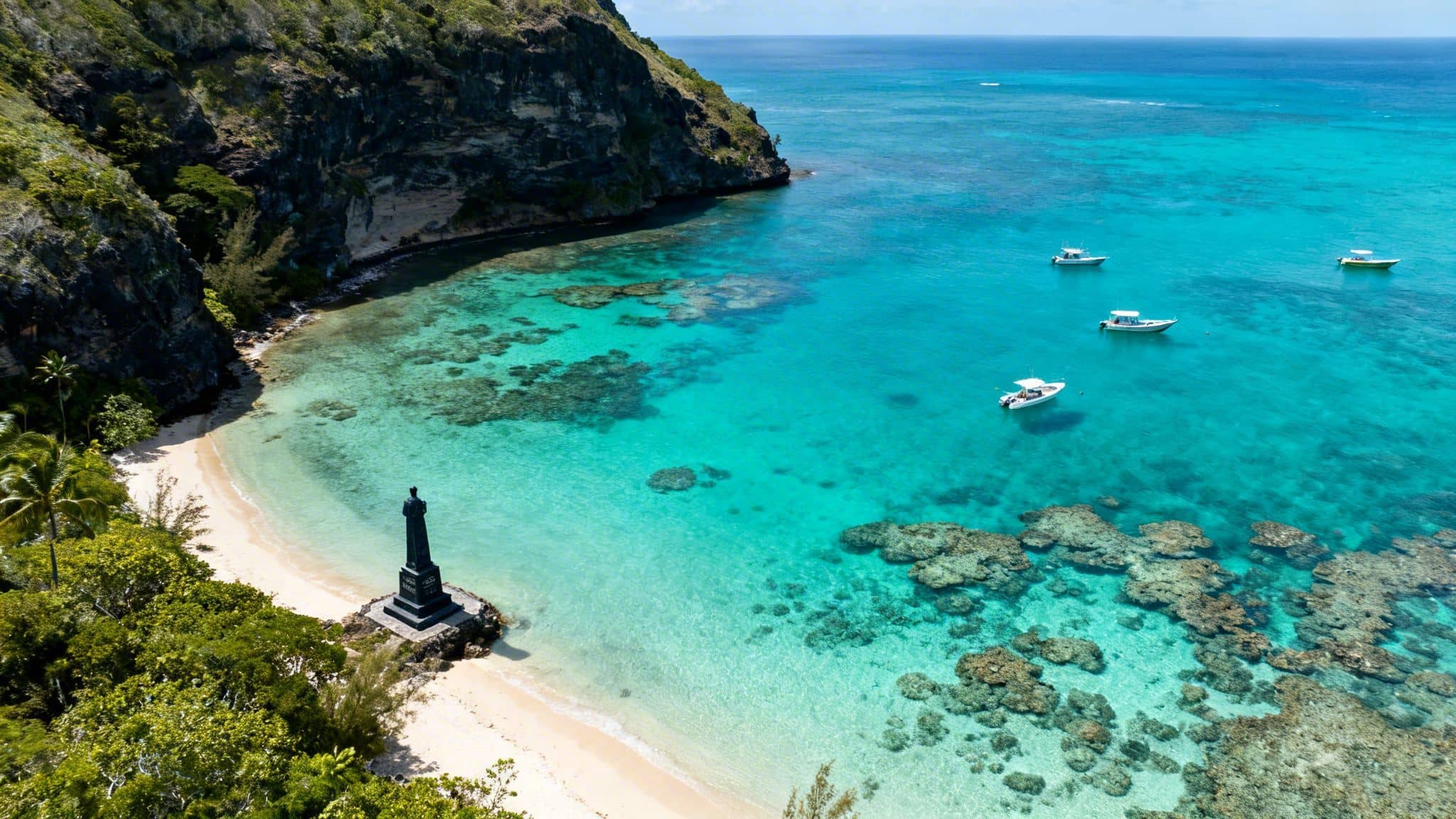 Aerial view of a vibrant tropical bay with clear turquoise water, sandy beach, green cliffs, monument, and boats.