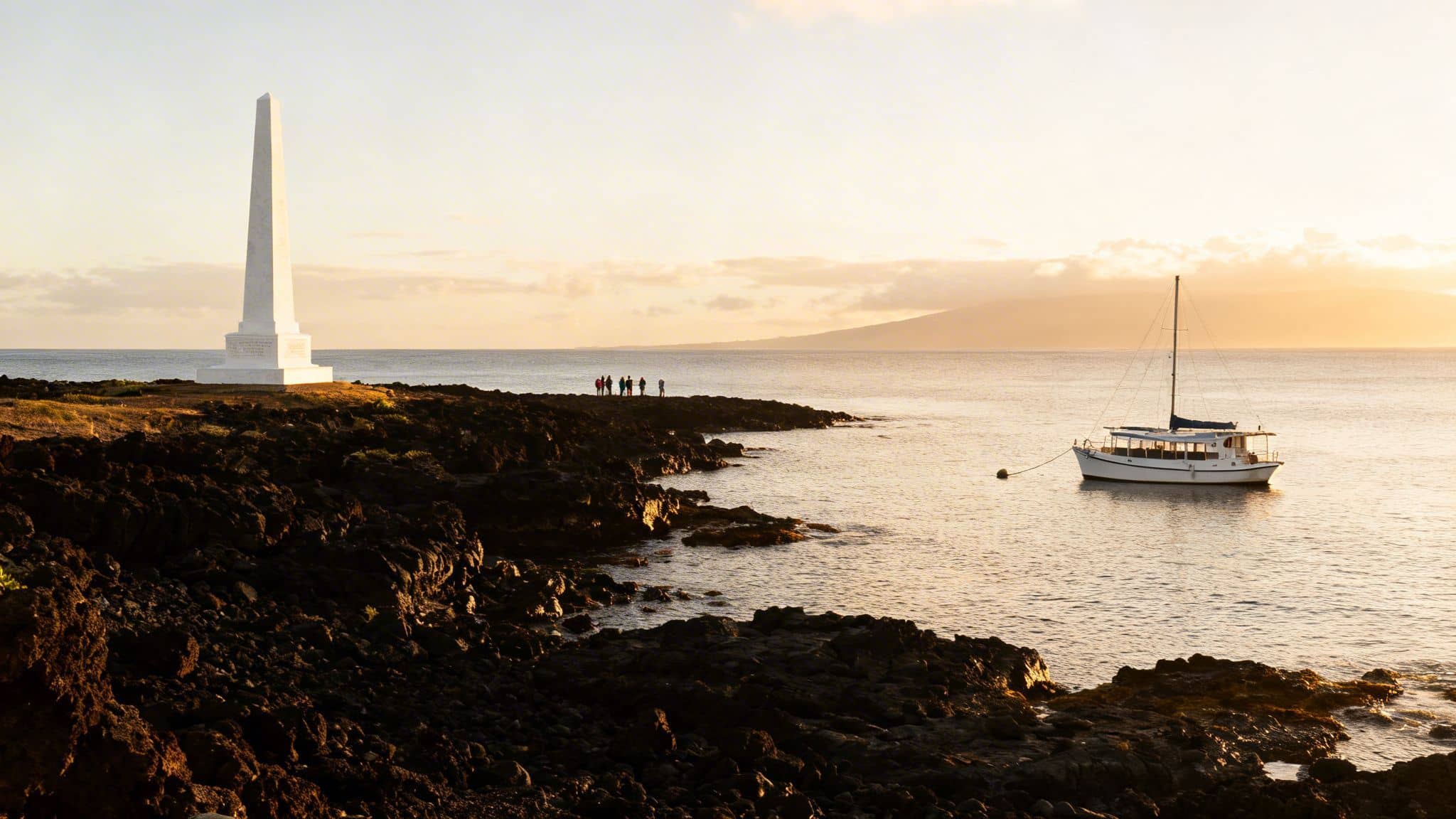 Golden sunset over Kealakekua Bay, showing Captain Cook Monument, people, and a boat.