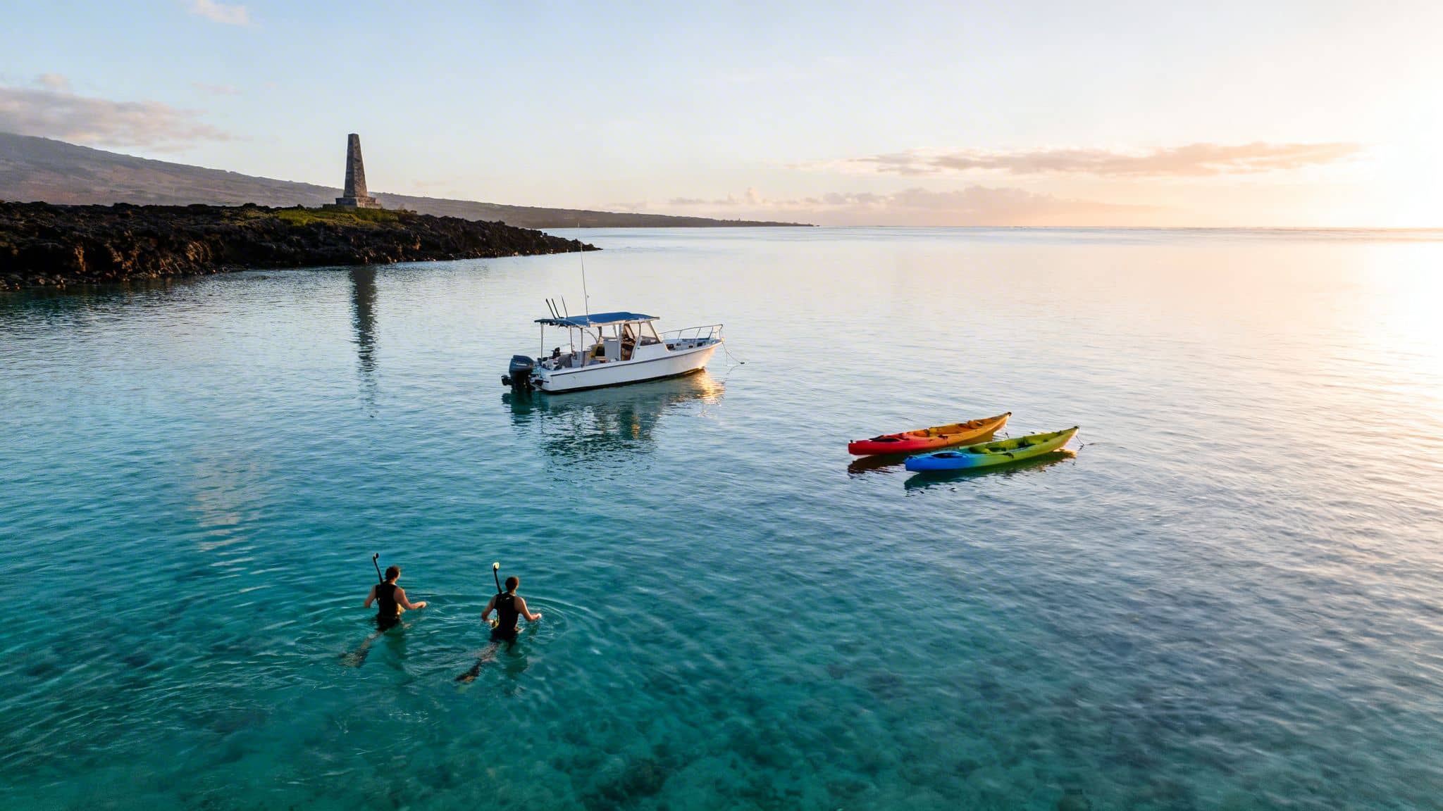 Aerial view of two snorkelers, a boat, and colorful kayaks in a beautiful bay with a historical monument.
