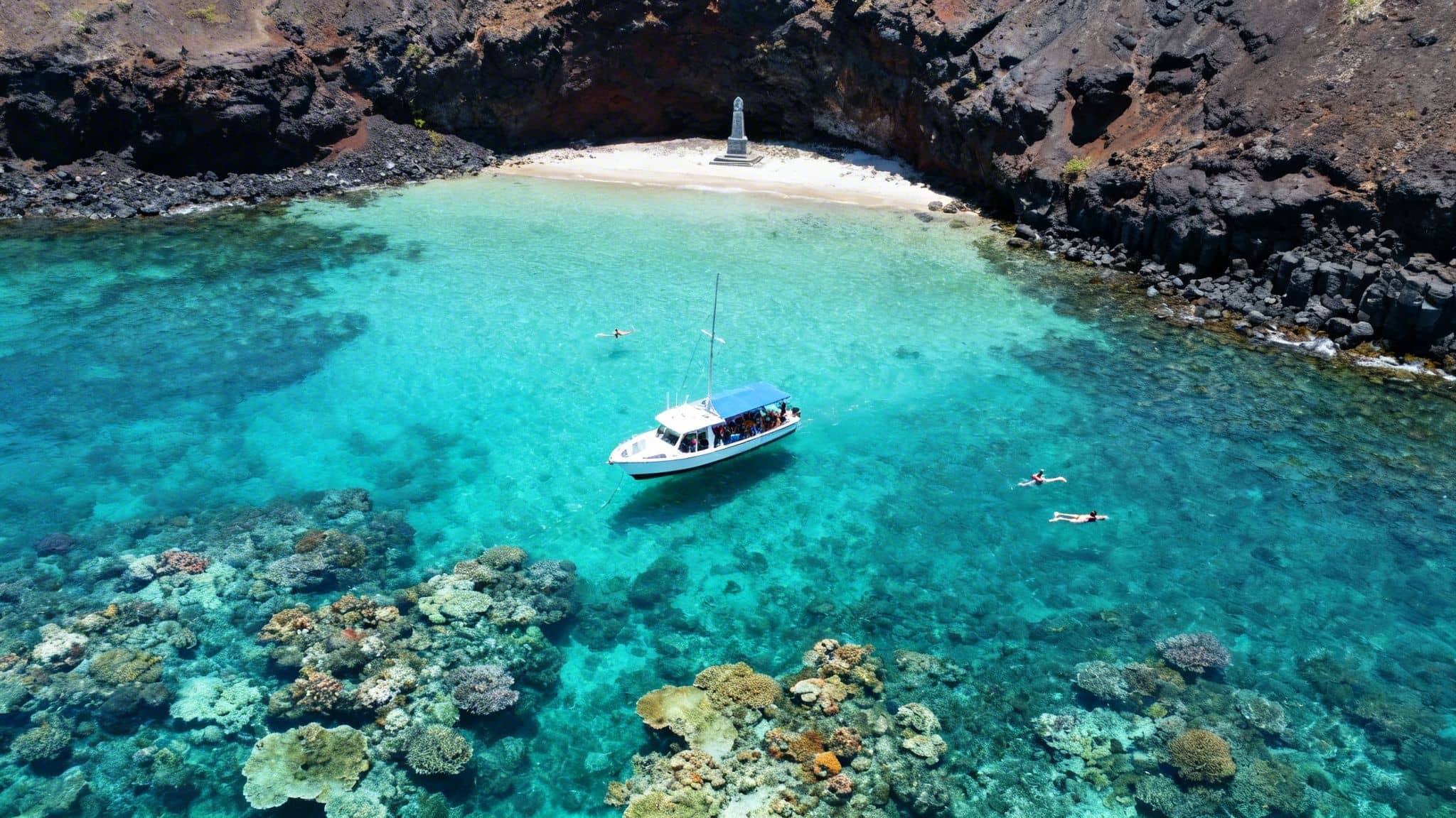 Aerial view of Kealakekua Bay with a boat, snorkelers, vibrant coral reefs, beach, and monument.