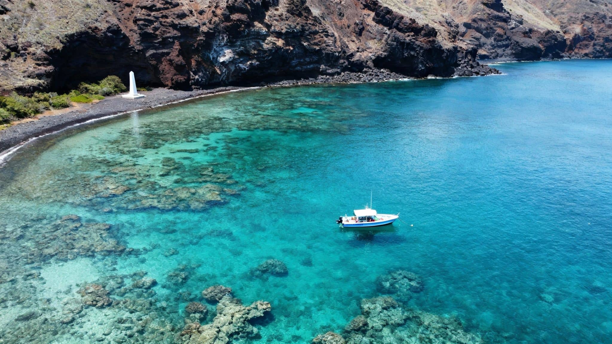 Aerial view of a white monument on a black pebble beach with a boat in clear blue water and coral.