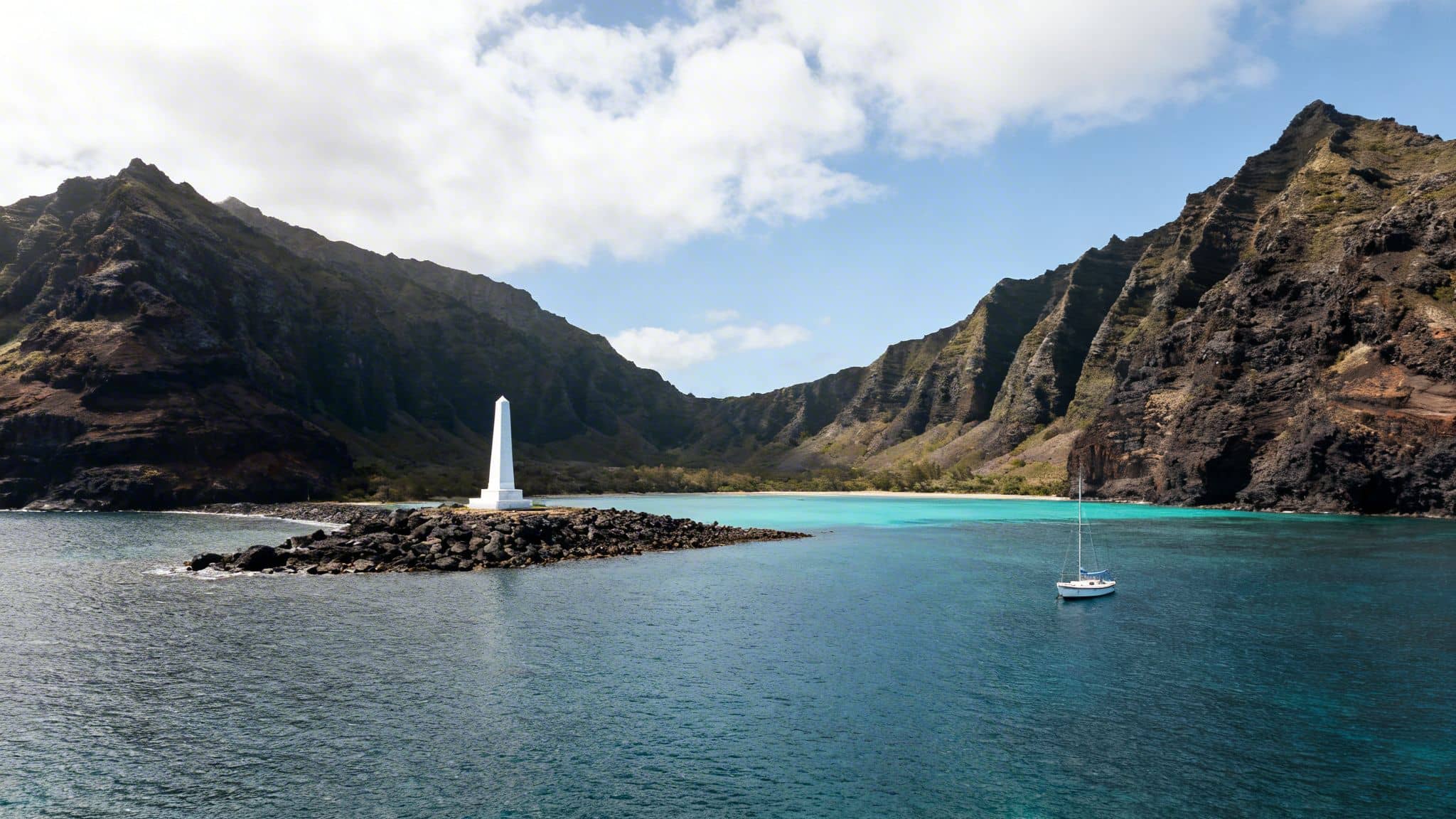A white monument on a rocky point overlooking turquoise water with a sailboat and lush mountains.