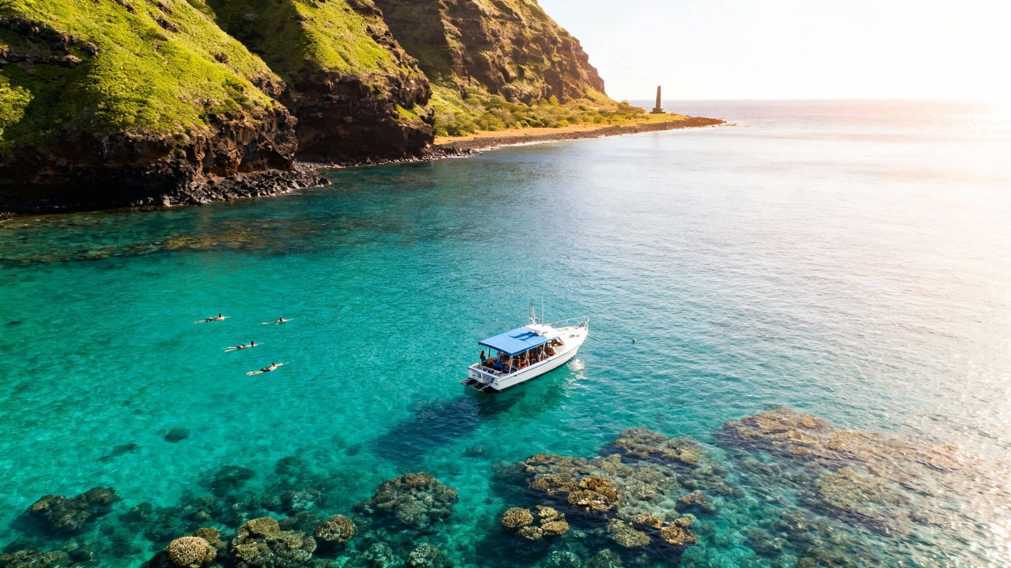 Aerial view of snorkelers, a boat, and coral reefs in turquoise ocean near a green cliffside with a distant lighthouse.