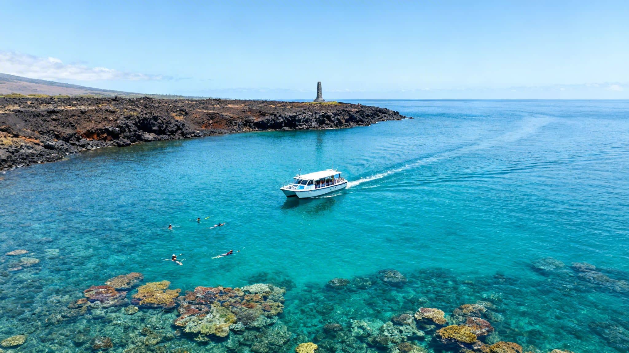 Aerial view of a boat and snorkelers in clear blue water near a volcanic coastline and monument.