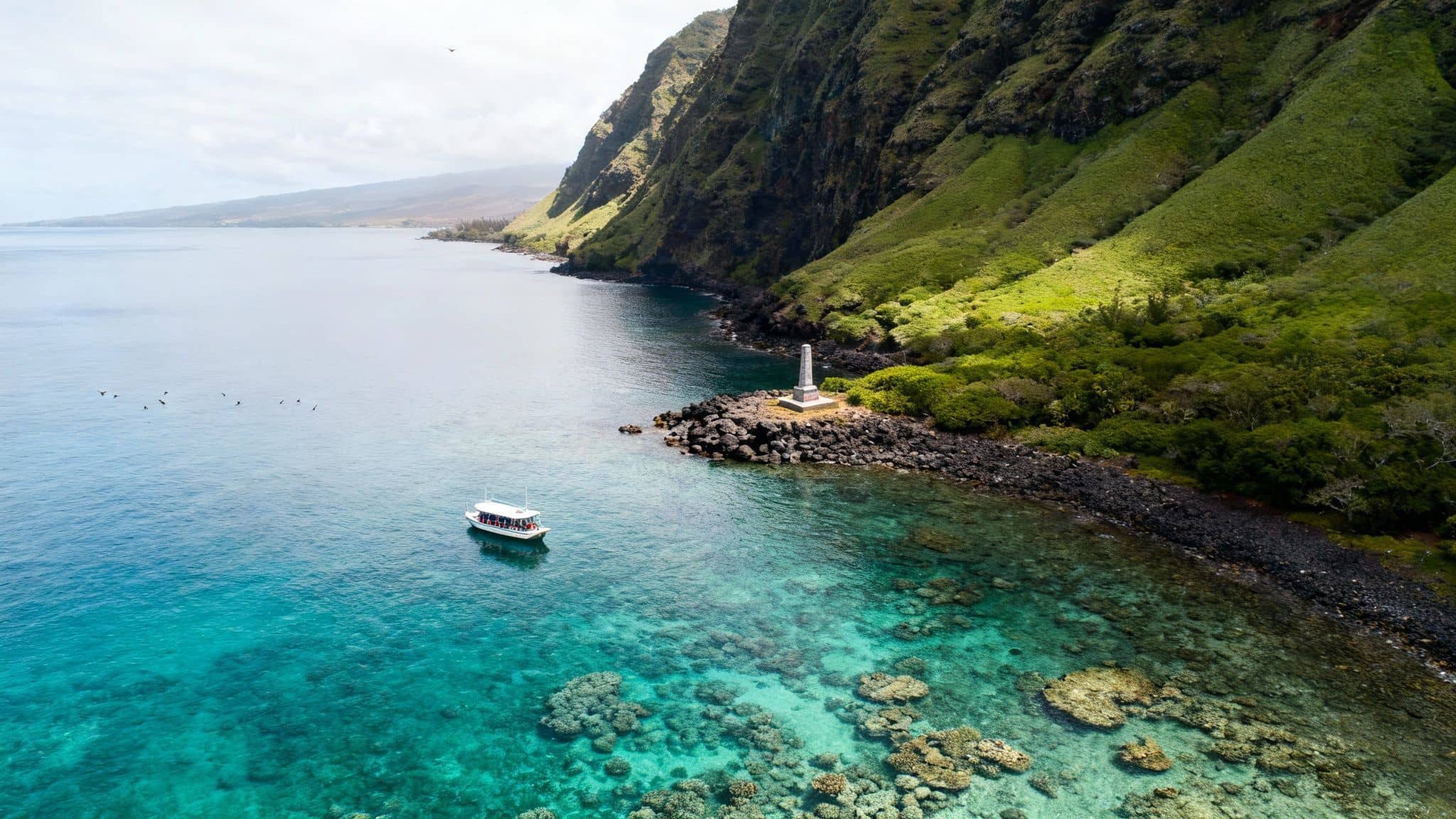Aerial view of a boat in clear blue water with visible coral reefs near a monument and green cliffs.