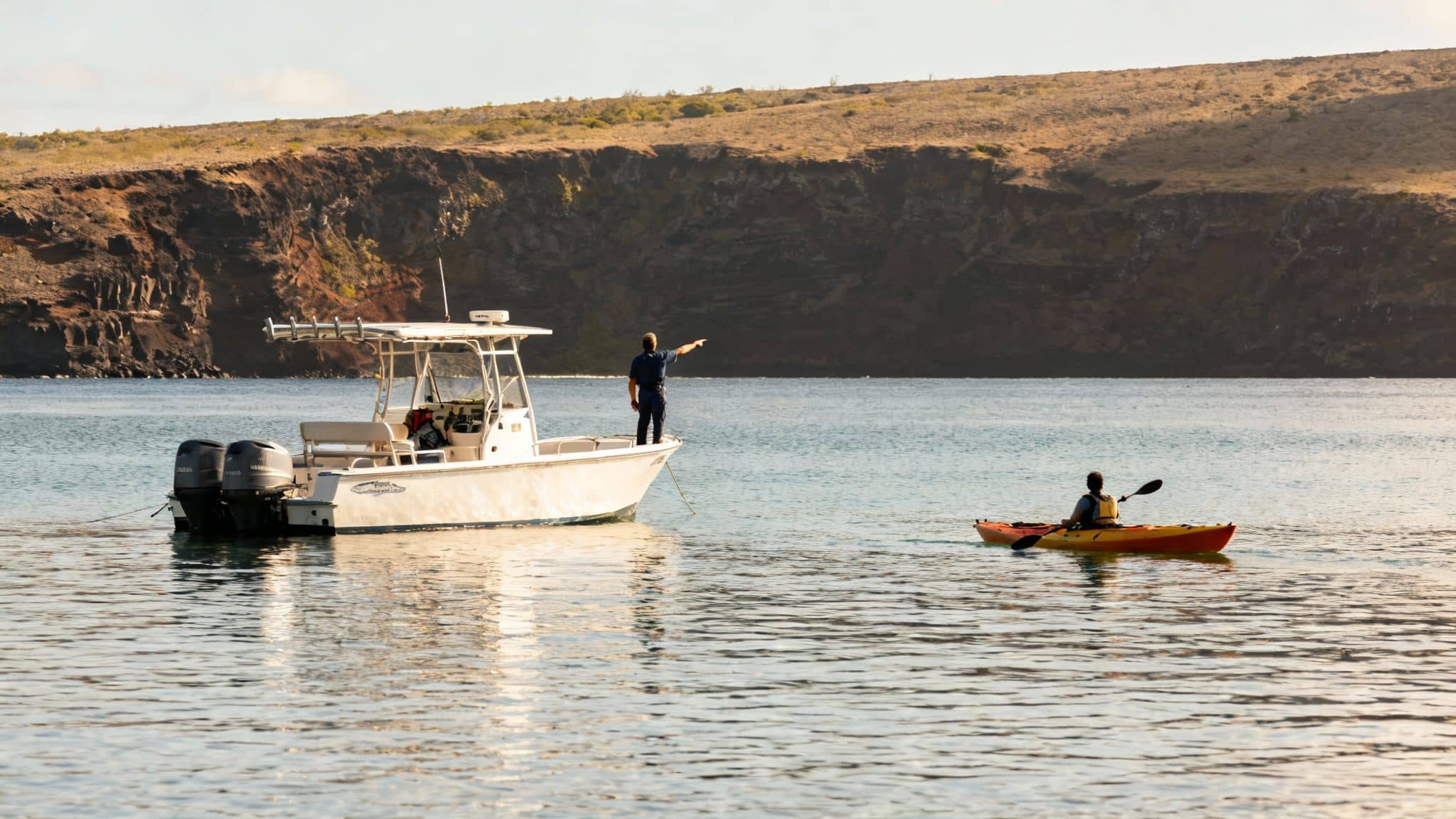 Two people in a yellow kayak paddling across the clear, turquoise water of Kealakekua Bay, with the Captain Cook Monument visible on the distant shore.