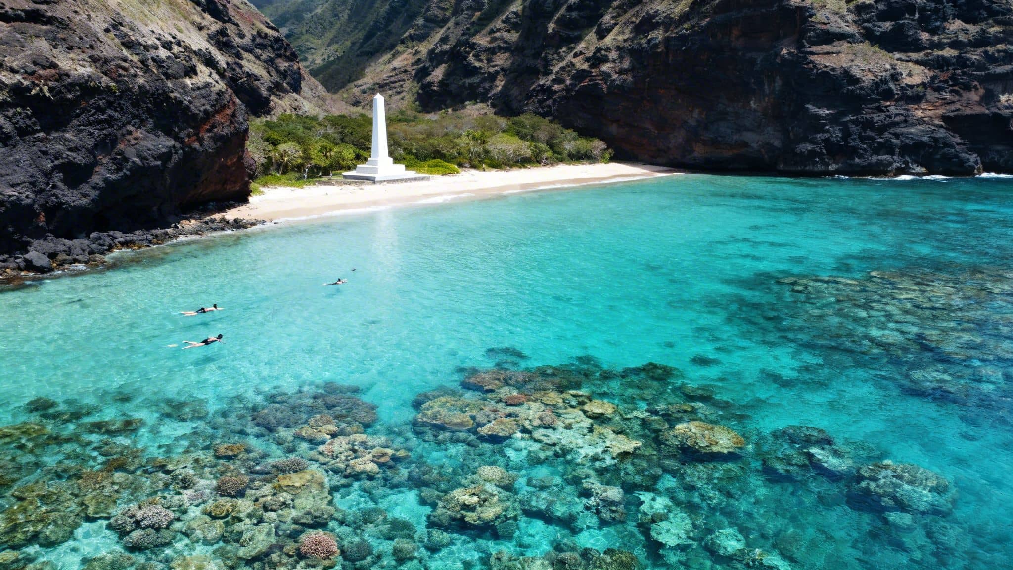 Vibrant turquoise bay with Captain Cook monument, lush cliffs, and snorkelers exploring coral.