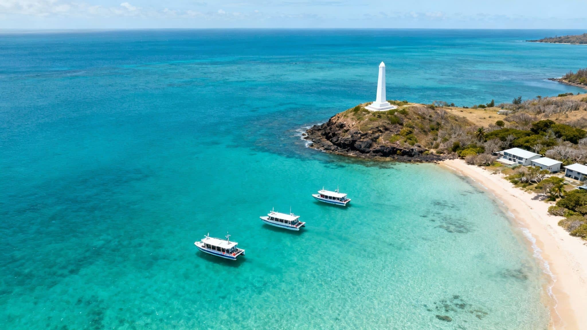 Three tour boats anchored in crystal clear turquoise water near white lighthouse and sandy beach