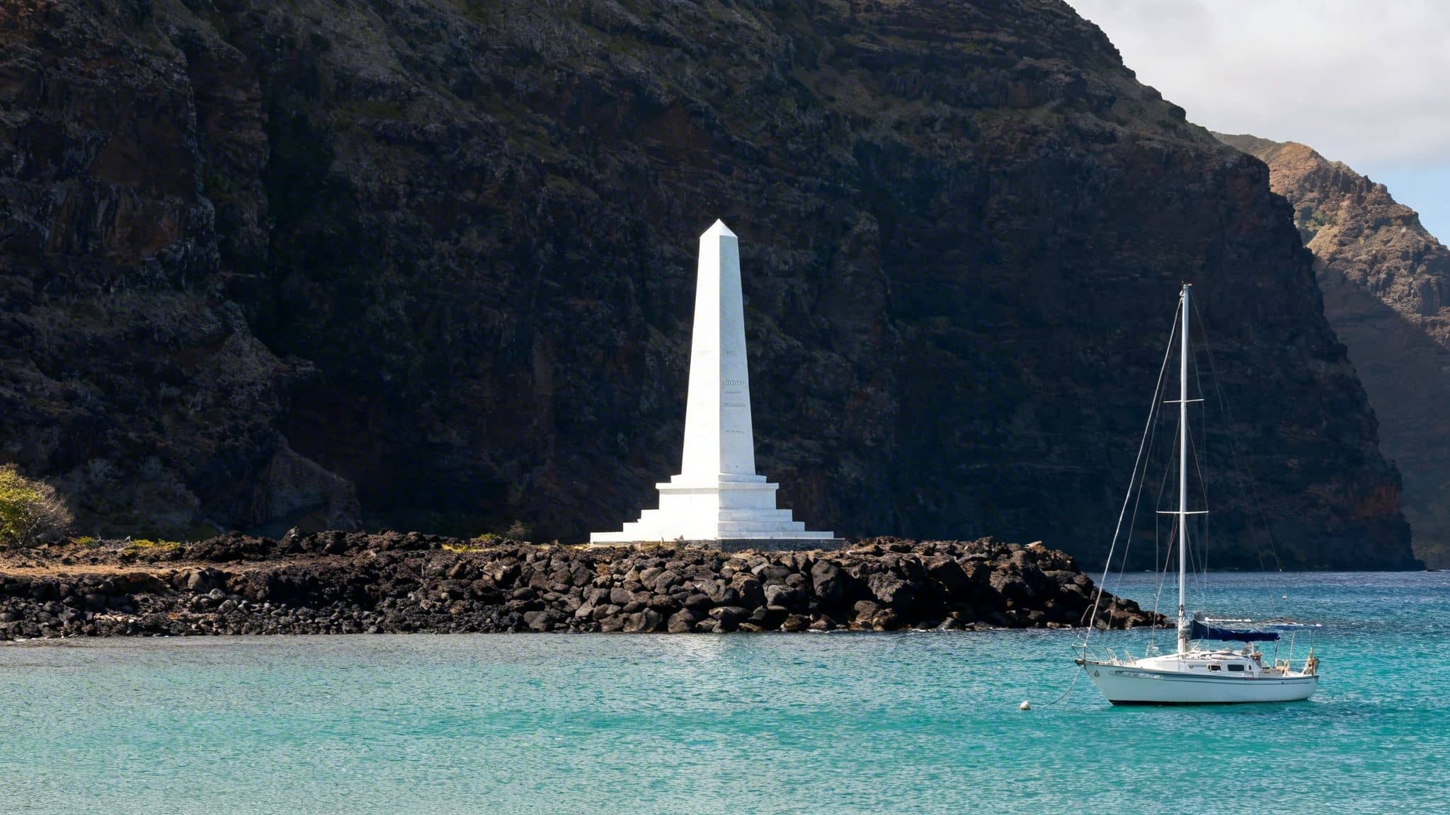 A white obelisk monument on a rocky shore, with a sailboat in turquoise water and dark cliffs.