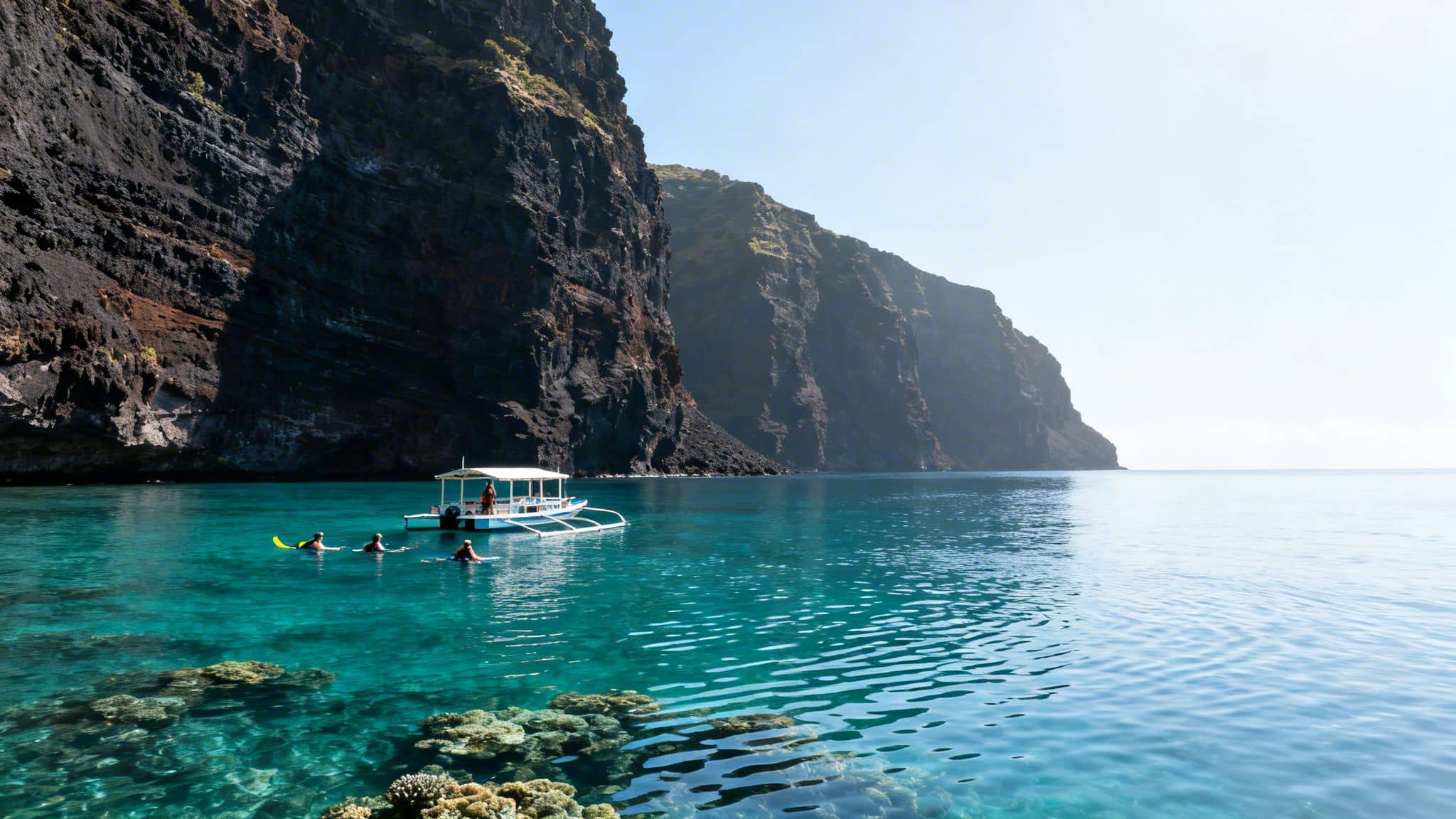 People snorkeling near a traditional boat in vibrant blue water alongside a dramatic volcanic cliff.
