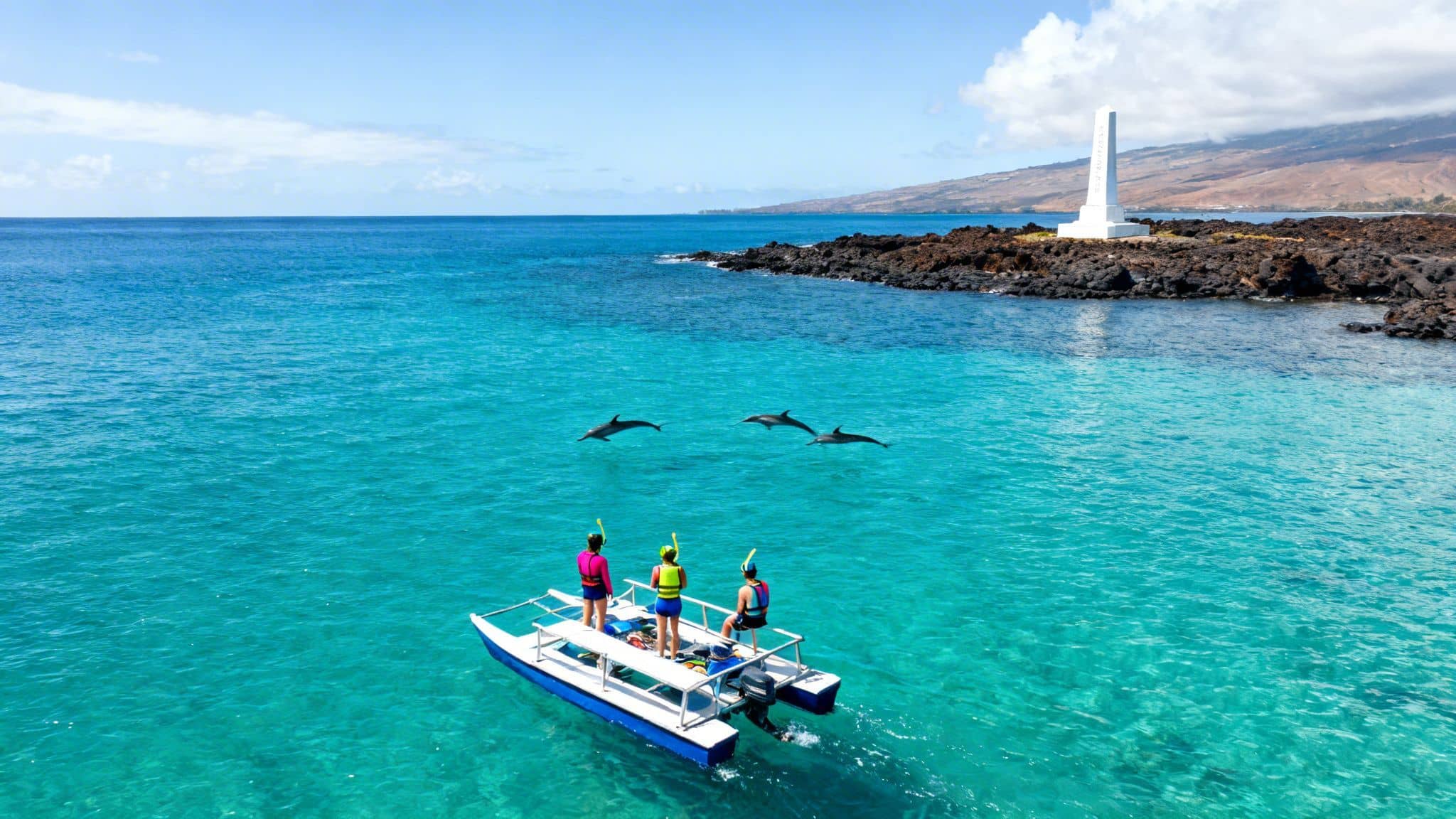 People on a snorkeling boat observe three dolphins swimming in clear turquoise water near a rocky Hawaiian coast.