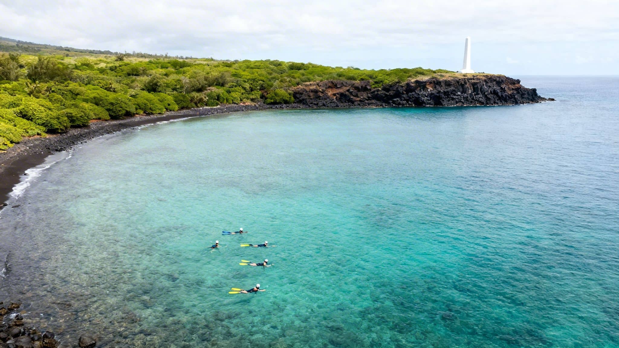 Aerial view of a black sand beach, clear turquoise water, snorkelers, and a white monument on a cliff.