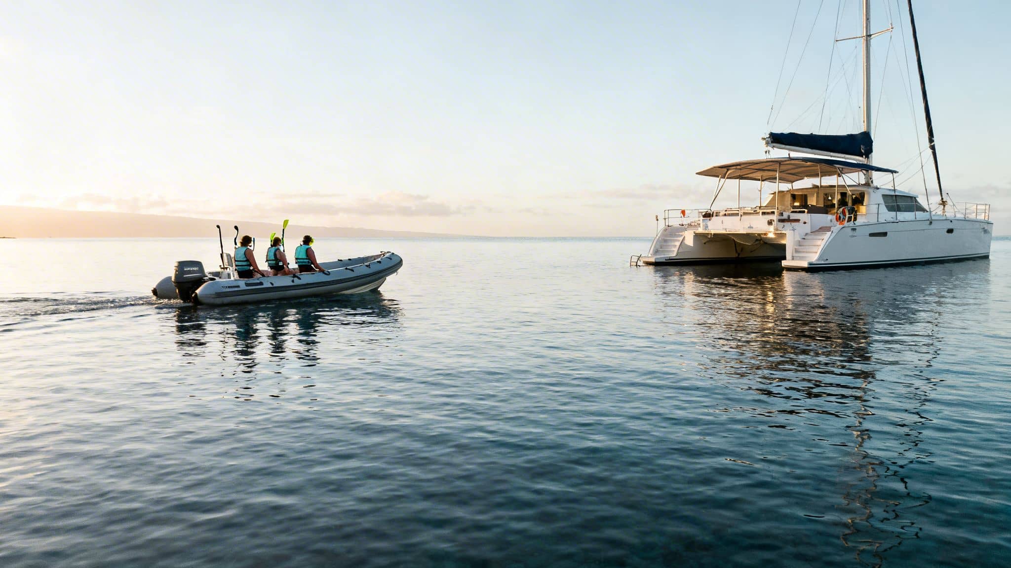Three people in a dinghy with snorkeling gear approach a large catamaran at sunset on calm ocean.