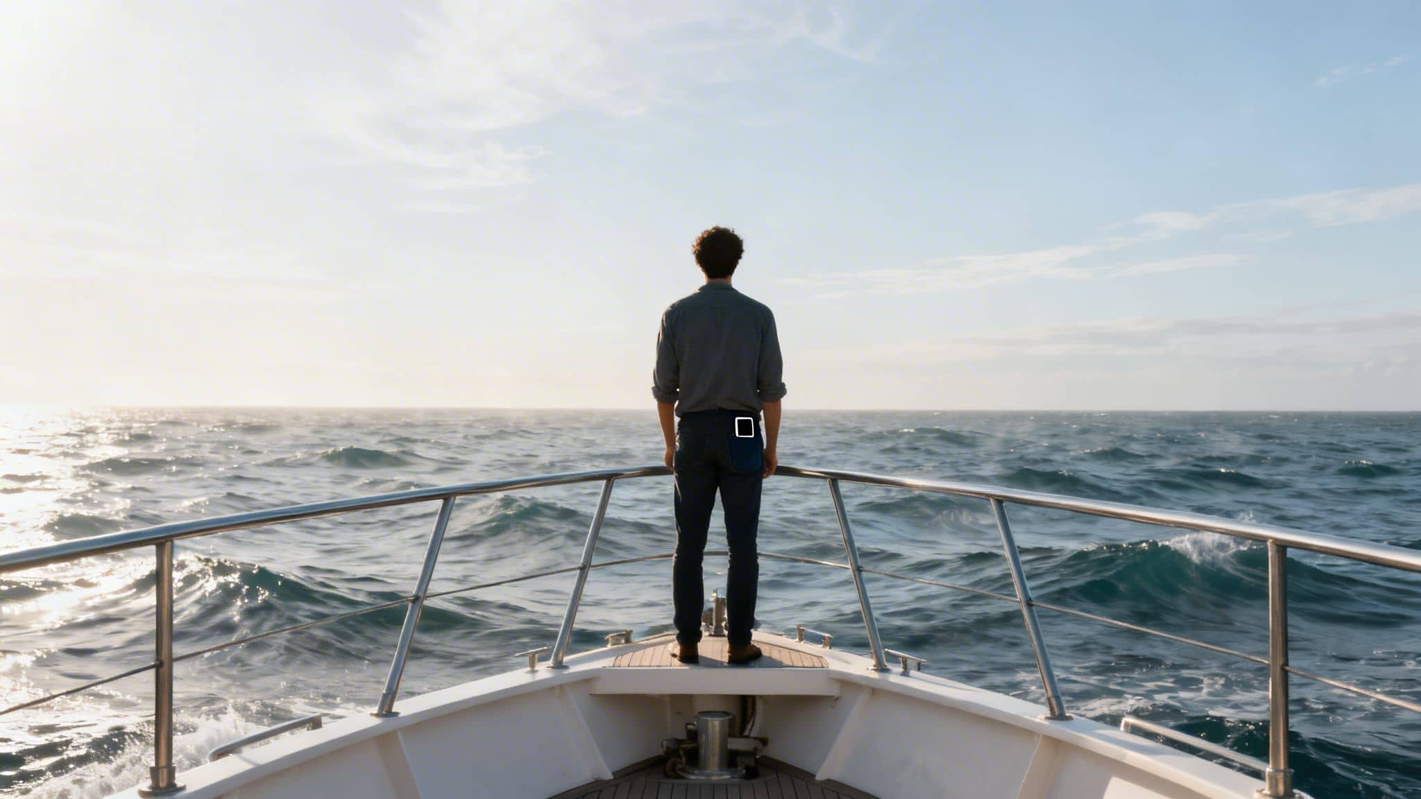 A man stands on the bow of a boat, gazing at the vast, sunlit ocean.