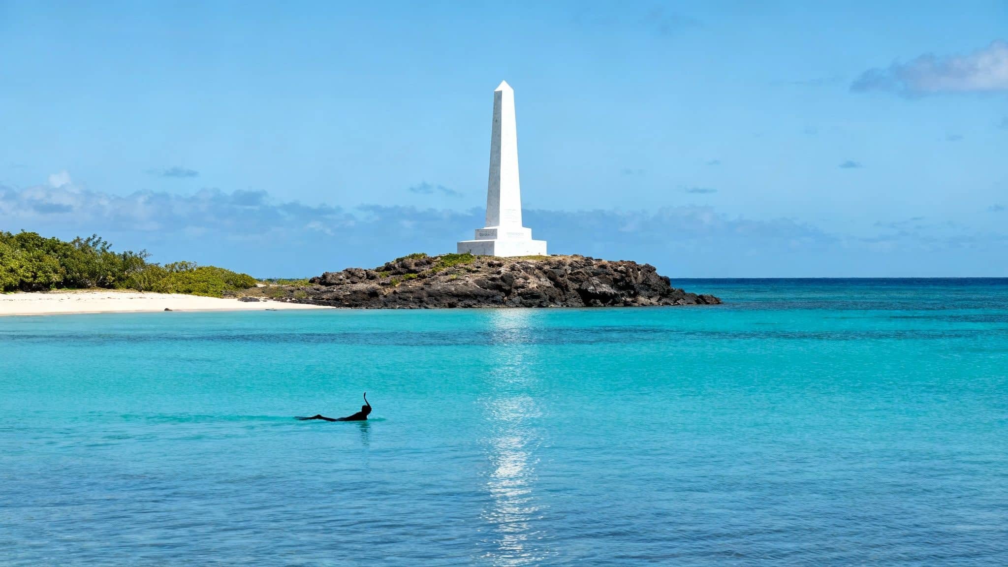 Snorkeler in tropical turquoise water near a white obelisk monument on a rocky island with a sandy beach.