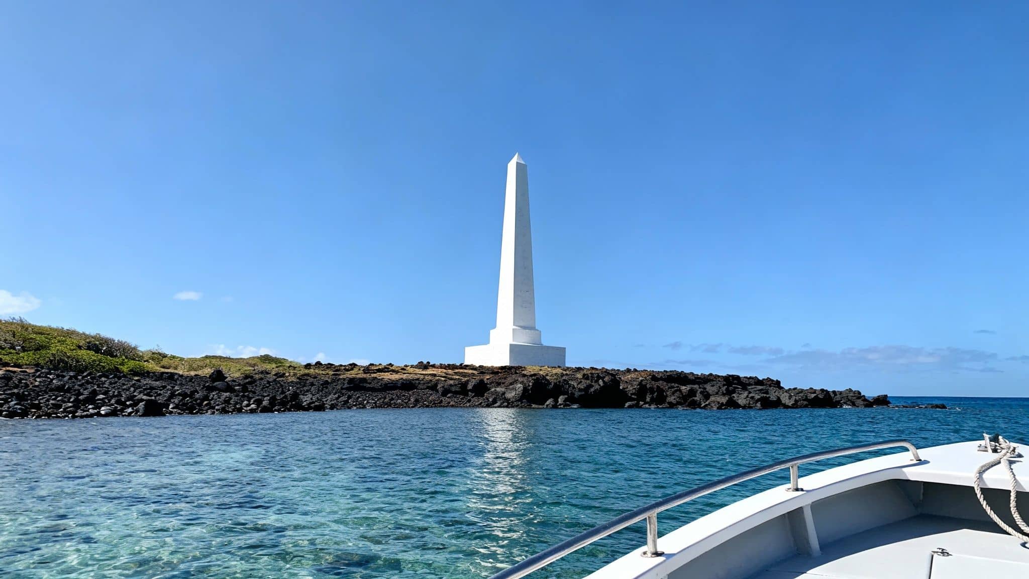 A striking white obelisk monument stands on a rocky island, viewed from a boat in clear blue ocean water.