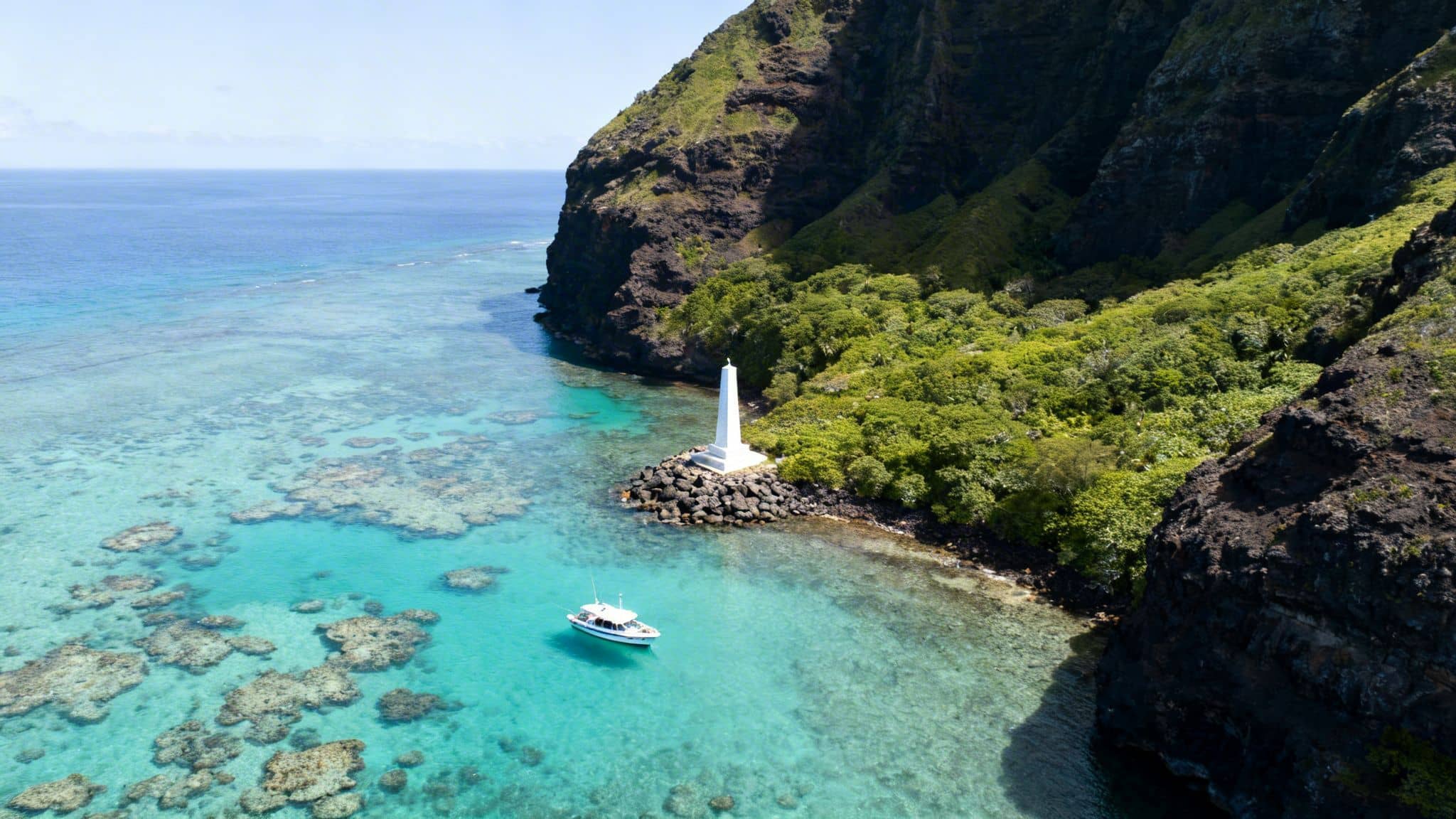 Aerial view of a white monument on a rocky Hawaiian coast with a boat in clear turquoise water.