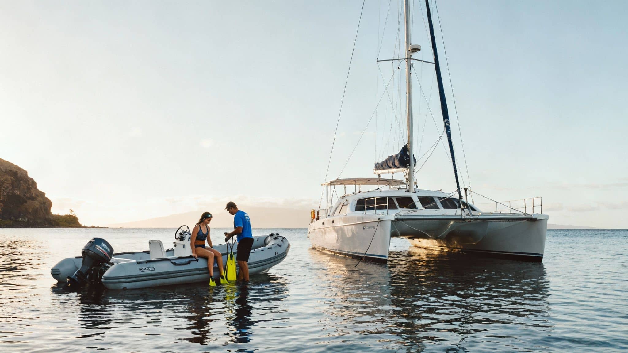 A couple prepares for snorkeling on a small boat with a catamaran sailboat anchored nearby.