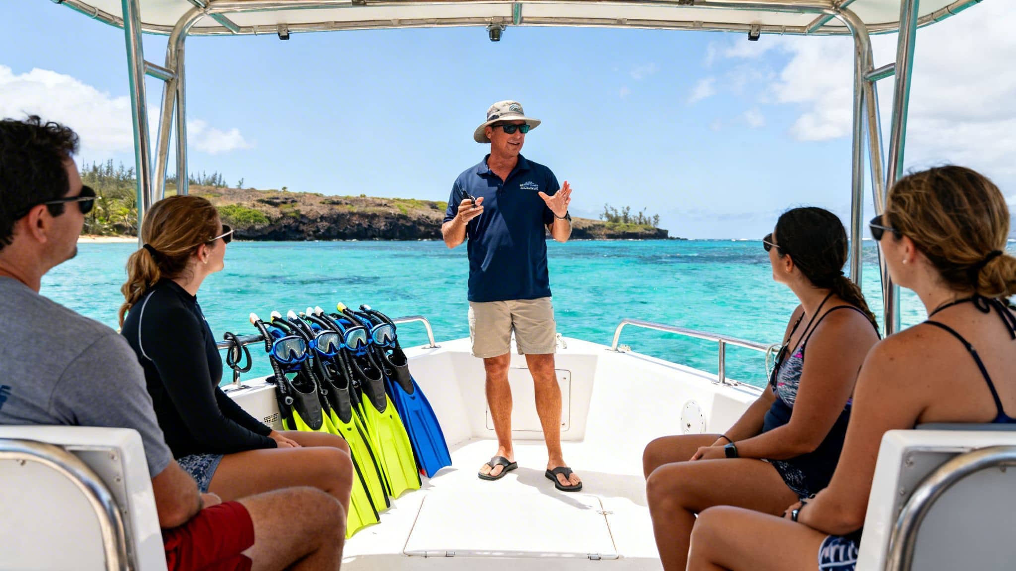 A tour guide on a boat speaks to a group of people, with snorkeling gear and clear ocean visible.