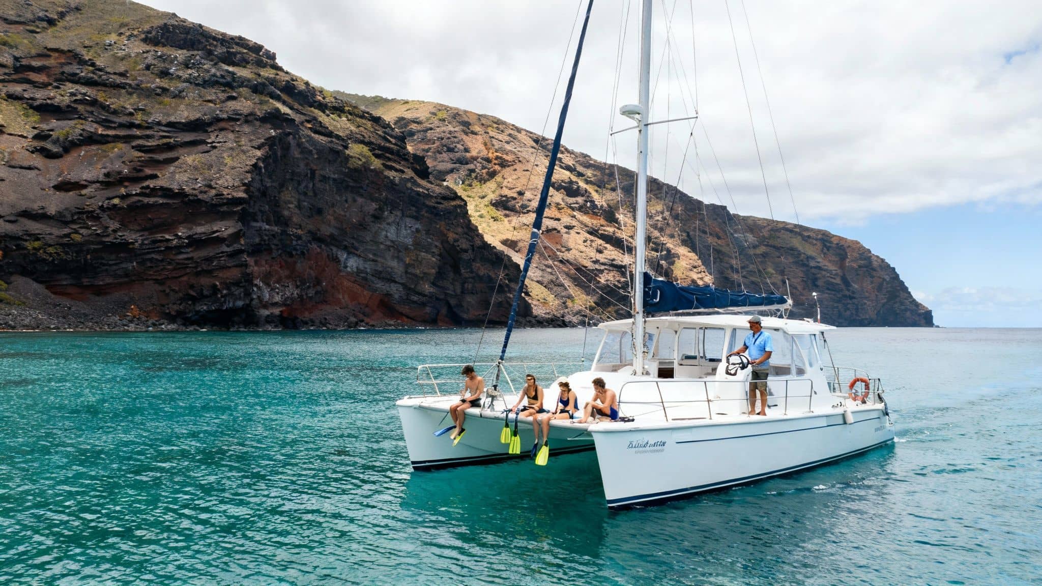 A white catamaran boat carrying people with snorkeling gear on turquoise water near dark rocky cliffs.