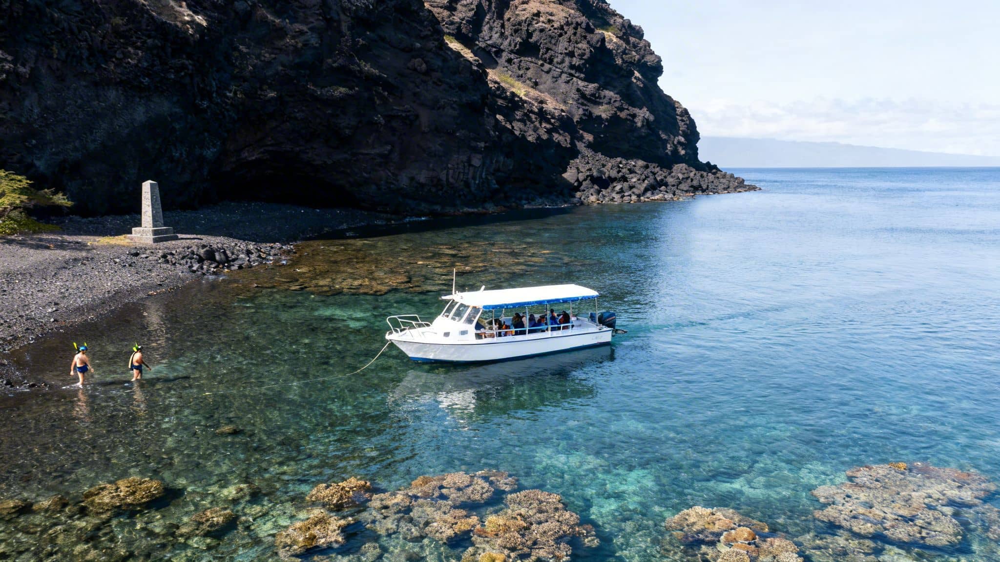 Snorkelers enter clear ocean water from a black pebble beach, near a boat and a monument.