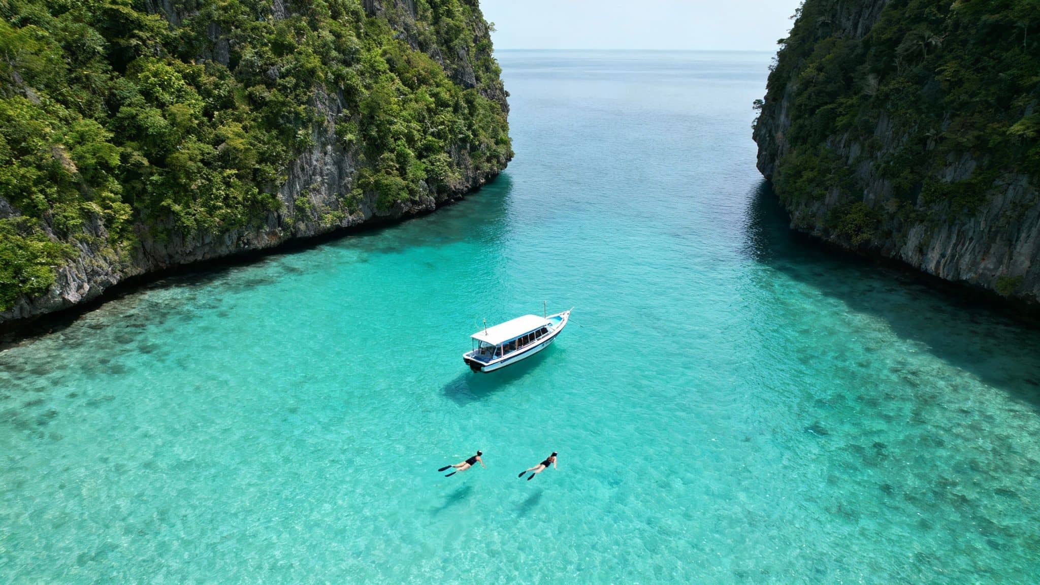 Snorkelers explore clear turquoise water near a boat in a beautiful tropical bay with lush green cliffs.