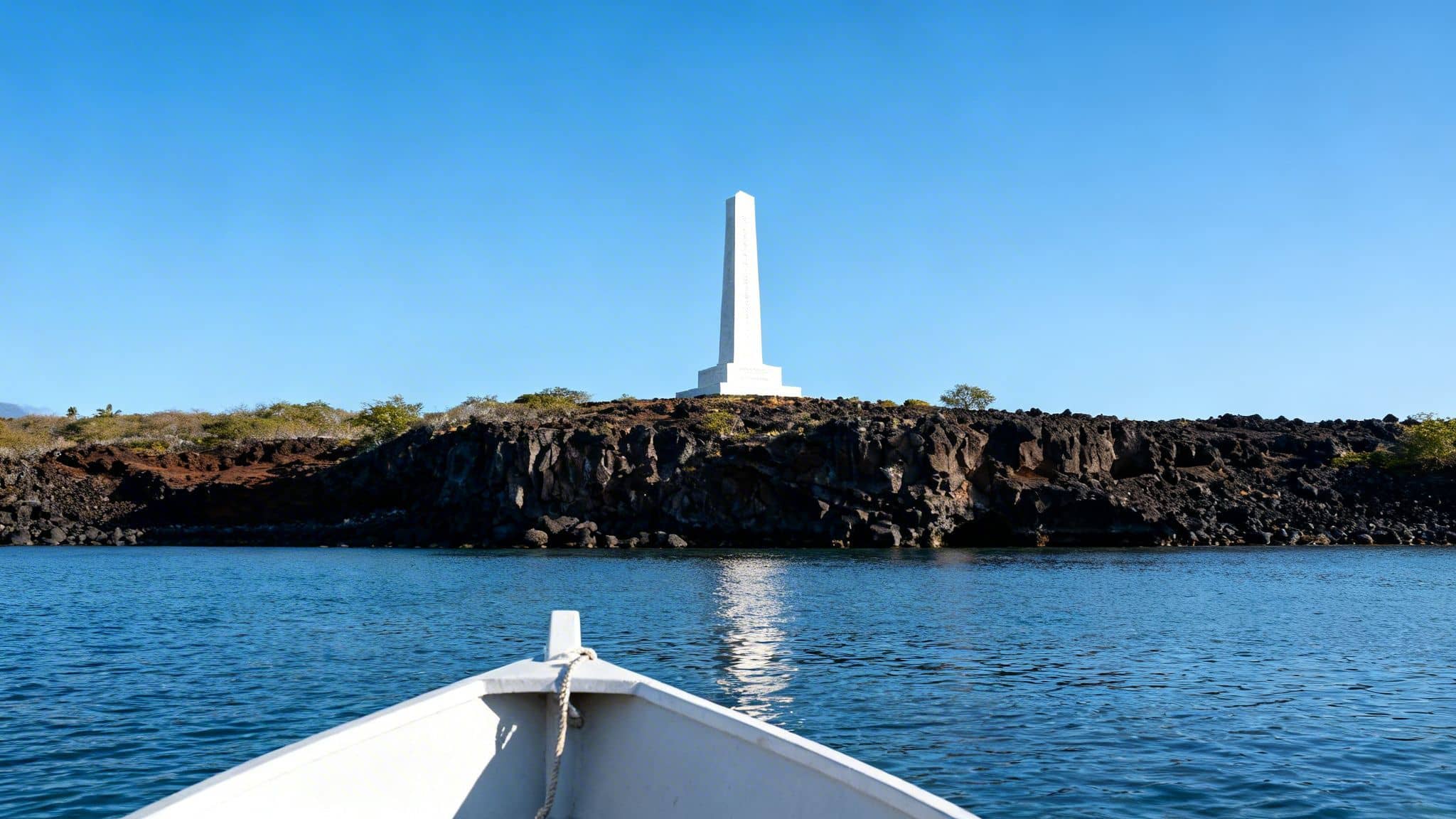 A white obelisk monument stands tall on a rocky island, seen from a boat in blue water.