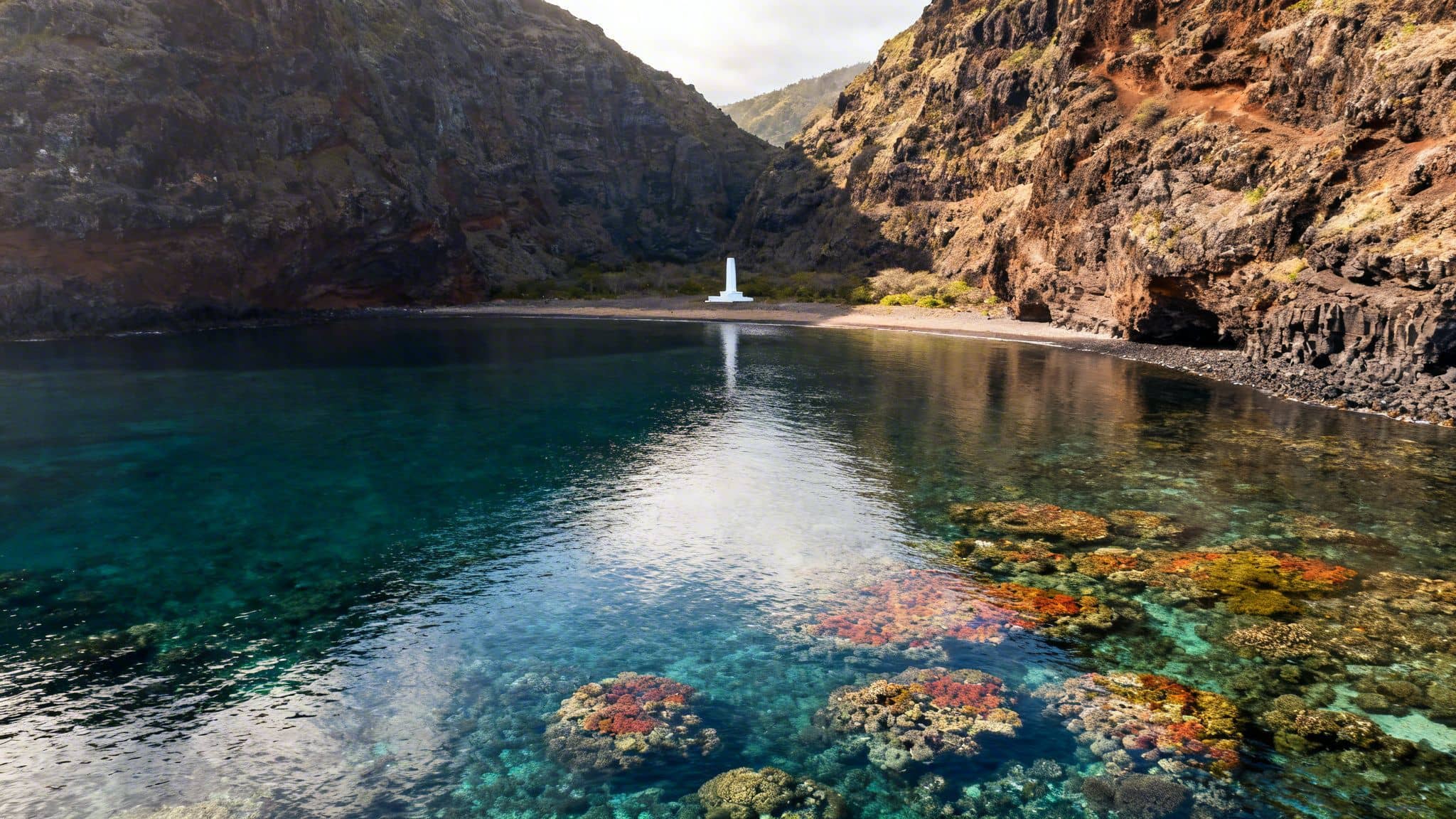 A scenic bay with colorful coral reefs in clear blue water, surrounded by steep cliffs and a white monument on a sandy beach.