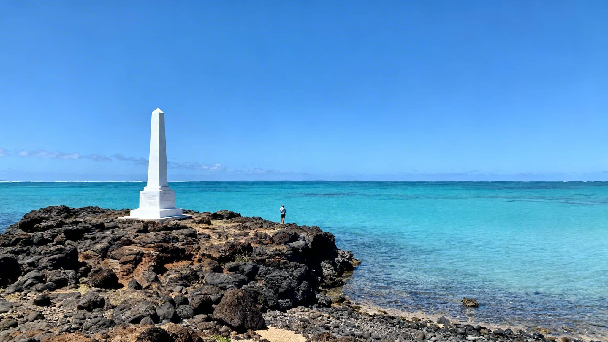 White obelisk monument on a volcanic rocky coastline overlooking vibrant turquoise ocean under a clear sky.