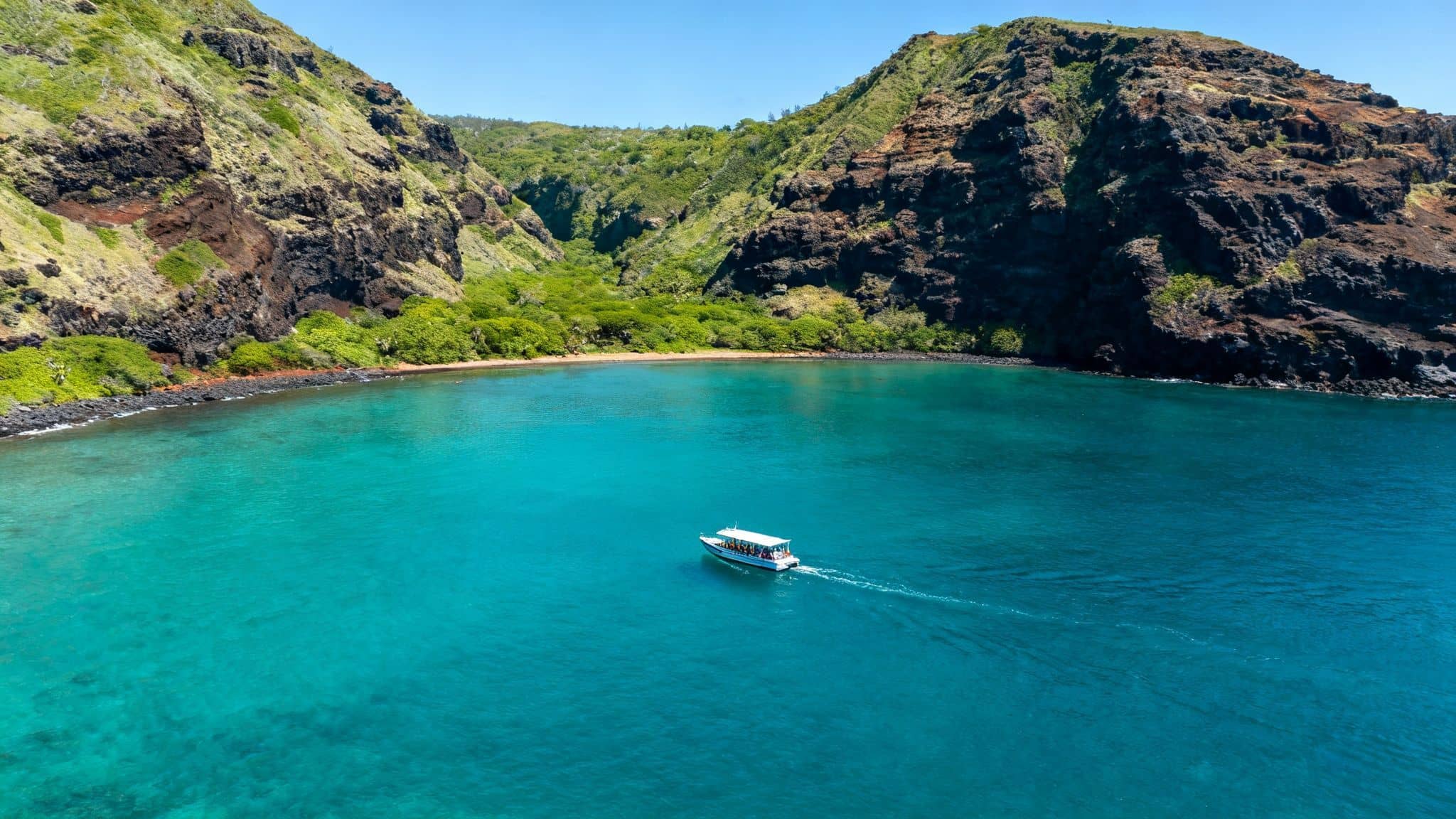 A small tour boat navigates the vibrant turquoise waters of a scenic tropical bay framed by lush green cliffs and a secluded beach.