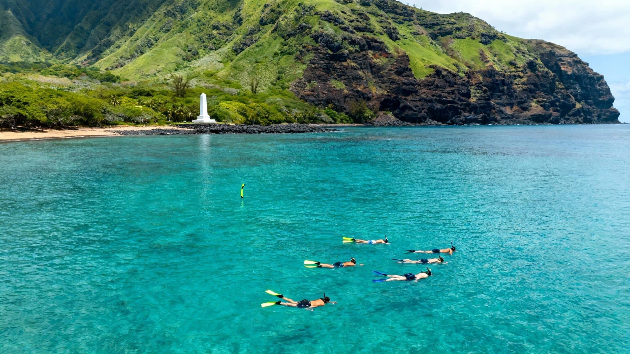 Group of people snorkeling in clear turquoise water near a tropical coastline with green mountains and a white monument.
