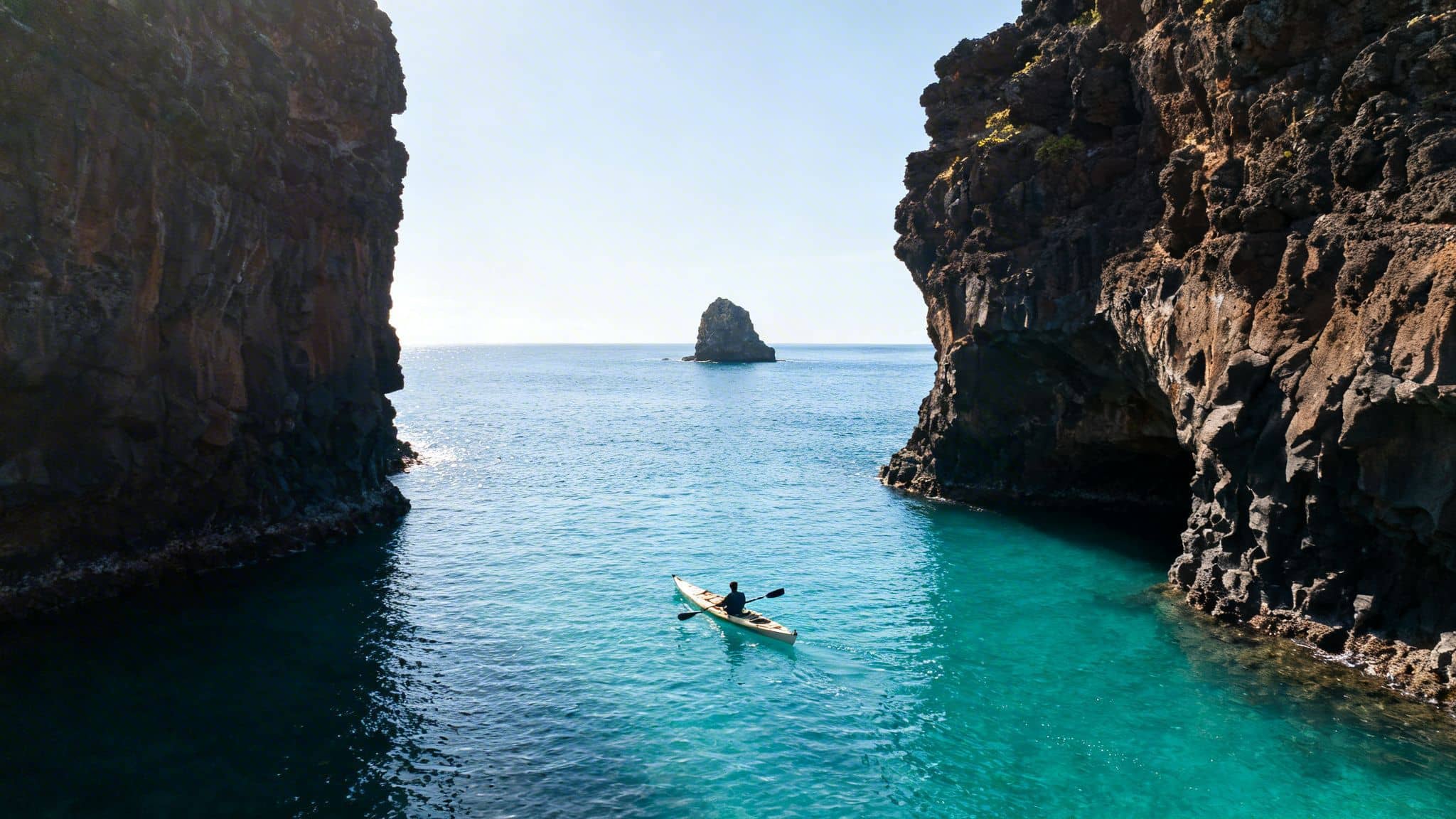 Kayaker paddles in stunning turquoise waters between dramatic sea cliffs, with an island in the distance.
