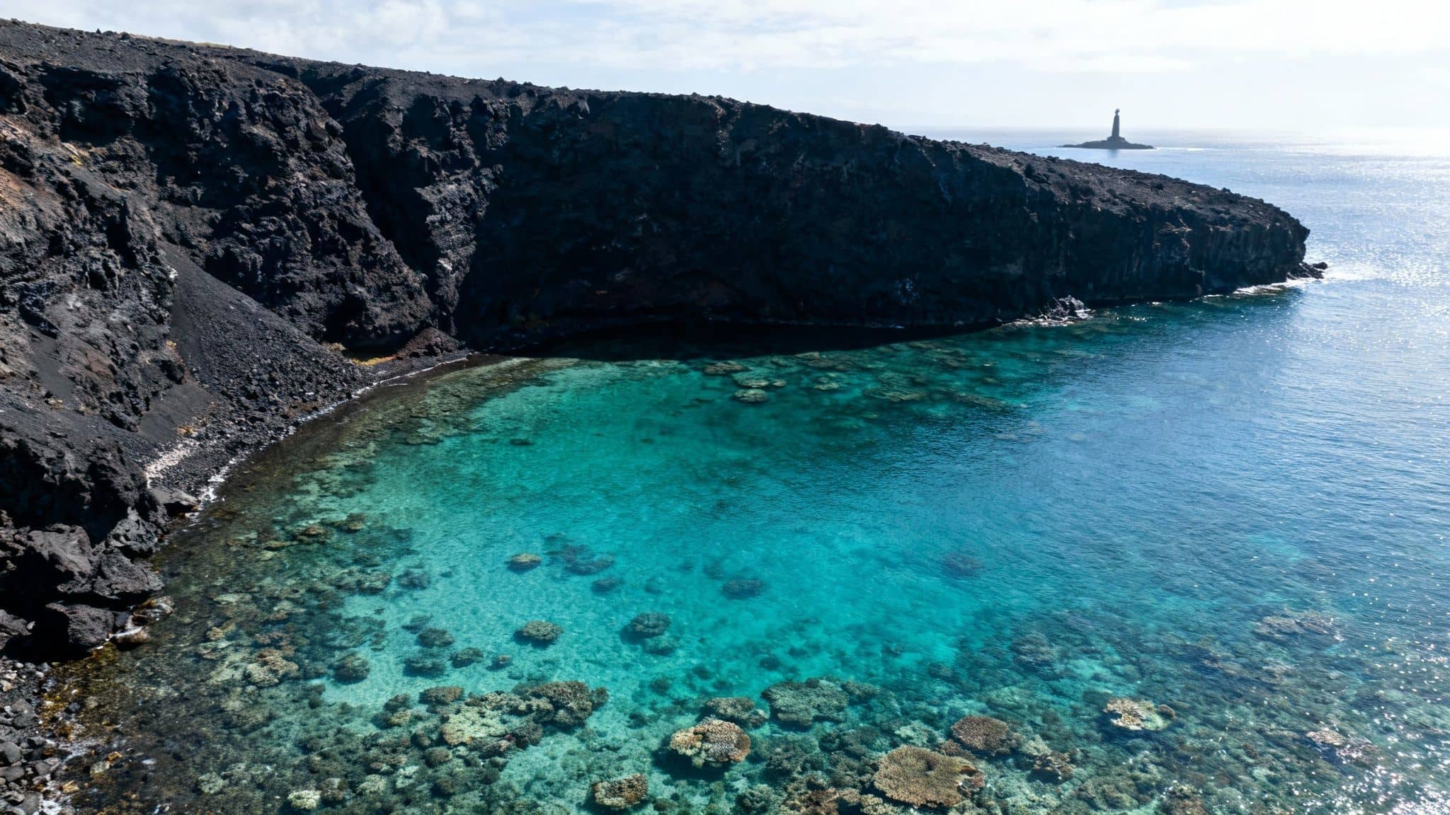 Aerial view of a rugged volcanic coastline with clear turquoise ocean water, revealing coral, and a distant lighthouse.