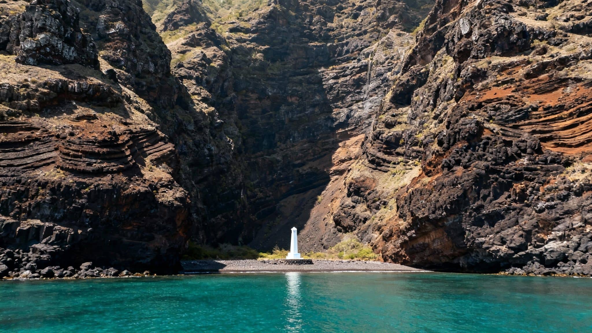 A narrow bay with towering rocky cliffs, turquoise water, and a white monument on a pebble beach.