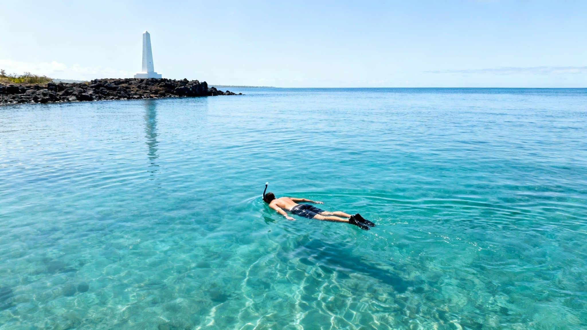 A person snorkeling in the clear turquoise ocean near a rocky island with a white monument.
