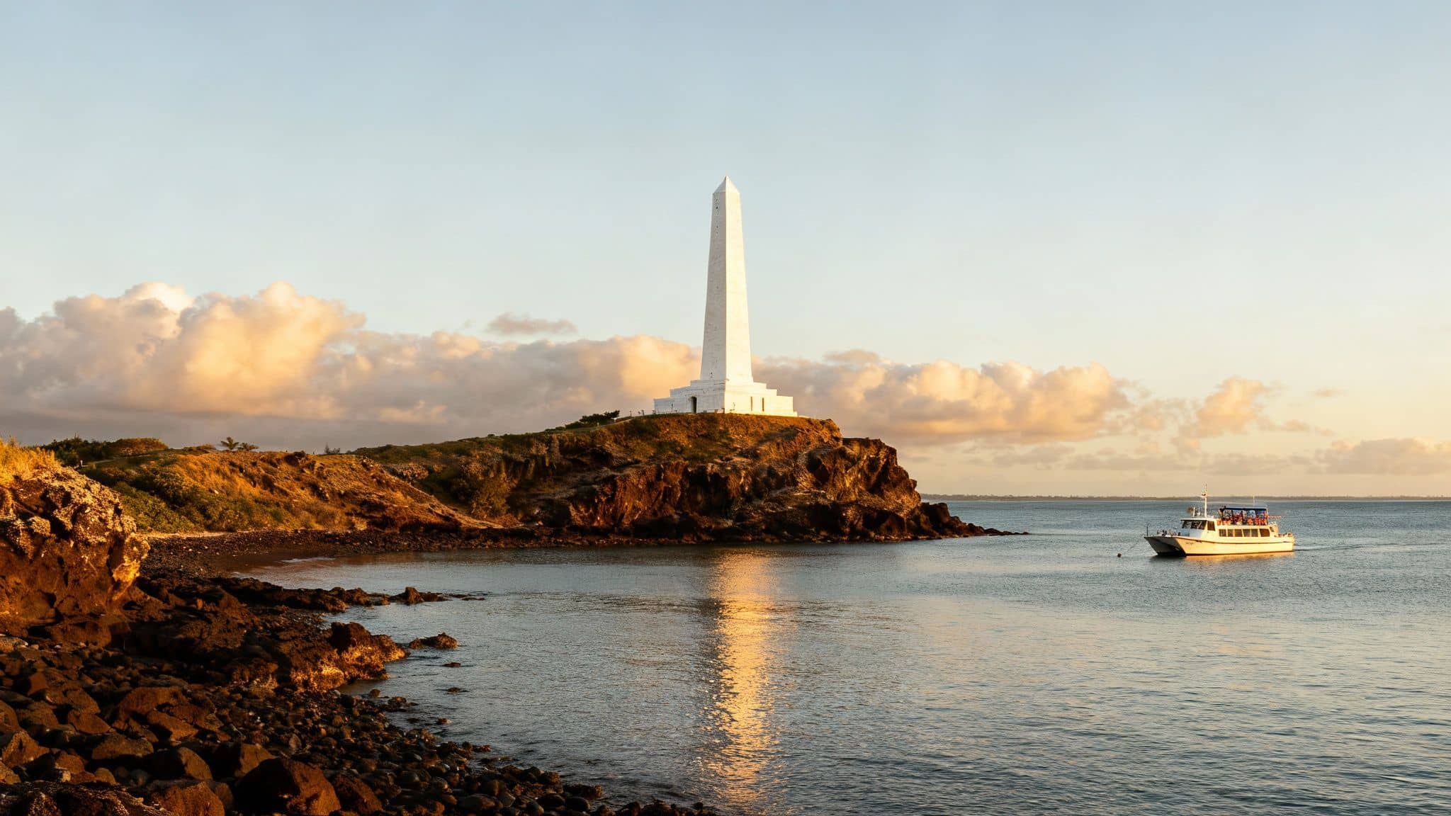 A white obelisk stands tall on a rocky cliff overlooking the ocean at sunrise, with a boat nearby.