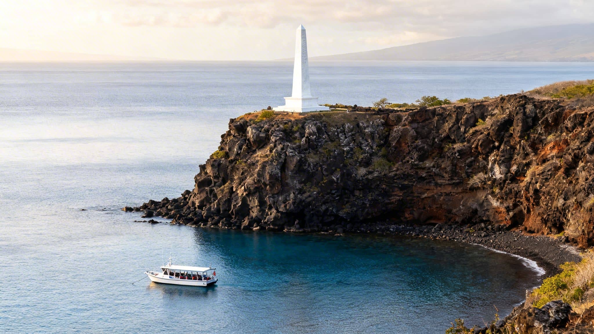 The white Captain Cook monument on a cliff overlooks Kealakekua Bay with a tour boat.