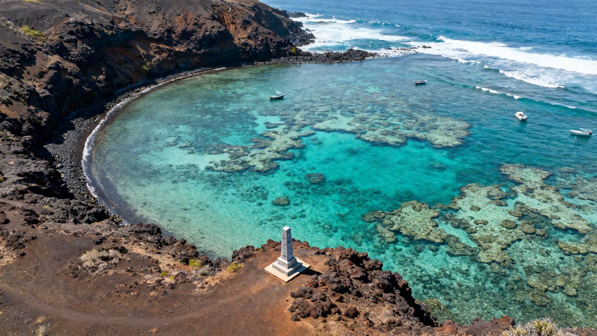 Aerial view of clear turquoise waters in a volcanic bay with coral reefs, boats, and a monument.