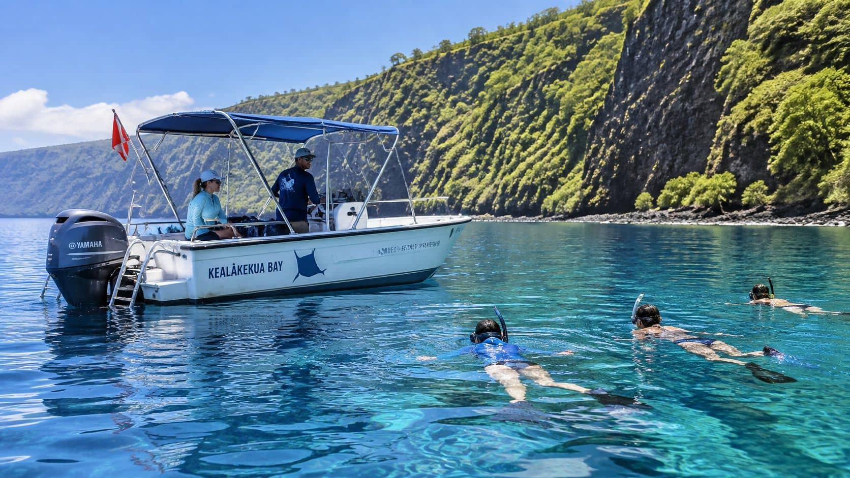 People snorkeling in the clear blue water of Kealakekua Bay near a boat with a cliff backdrop.