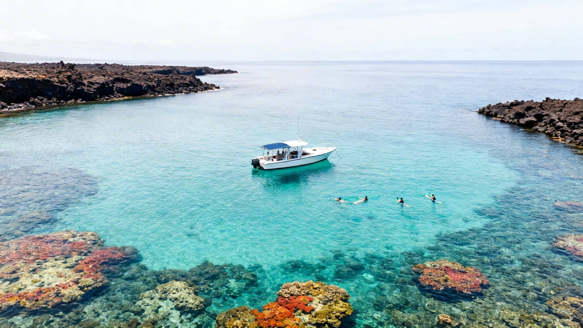 Aerial view of a boat anchored in a clear turquoise bay with people snorkeling over a vibrant coral reef.