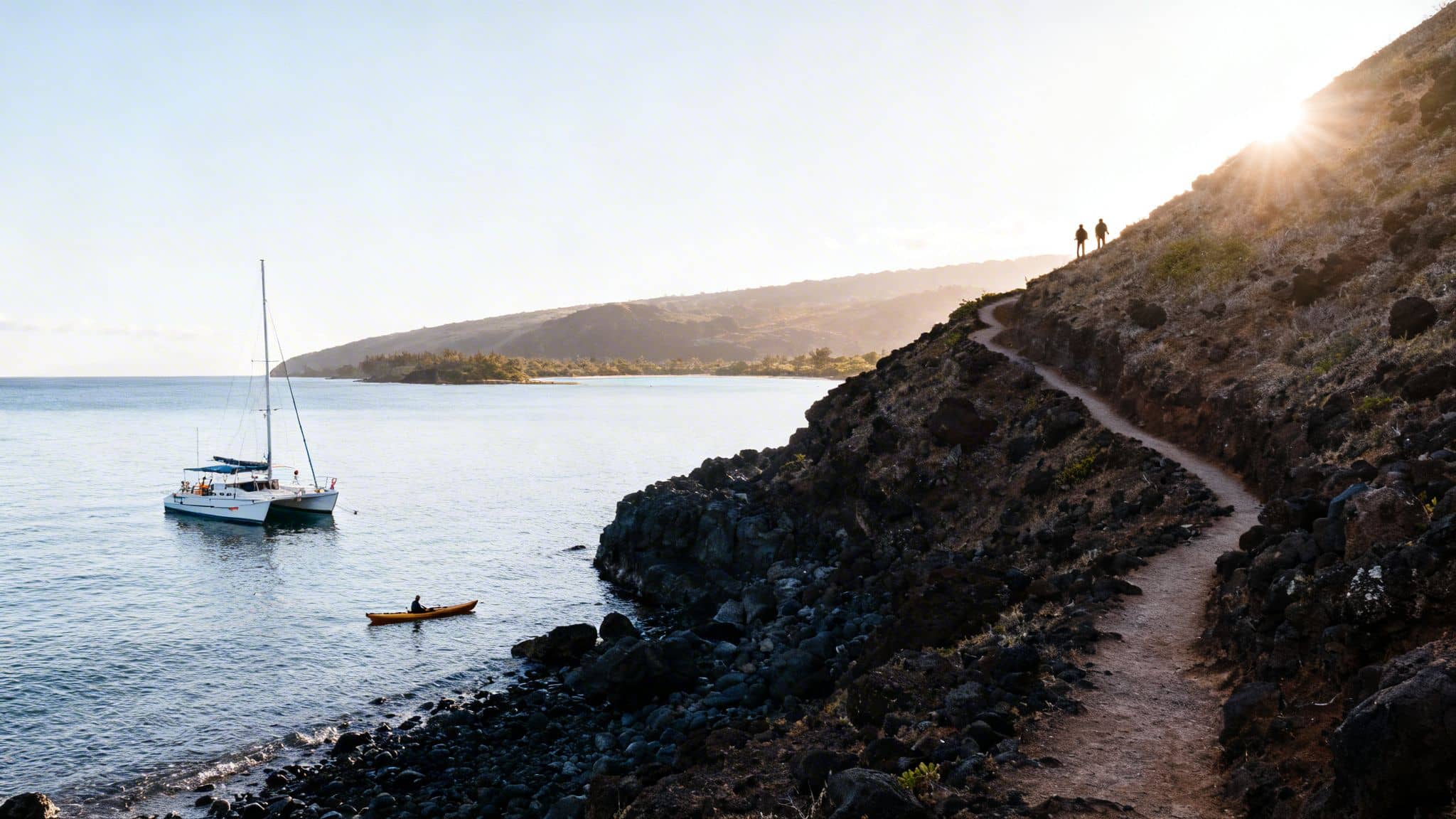 Scenic coastal view with a catamaran, kayaker, and hikers on a winding path at sunset.