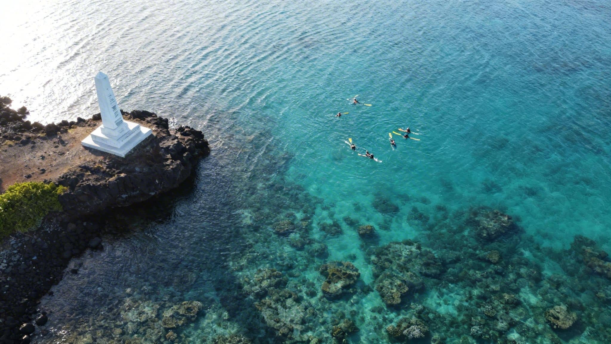 Aerial view of the white Captain Cook Monument on a rocky point, with people snorkeling nearby.