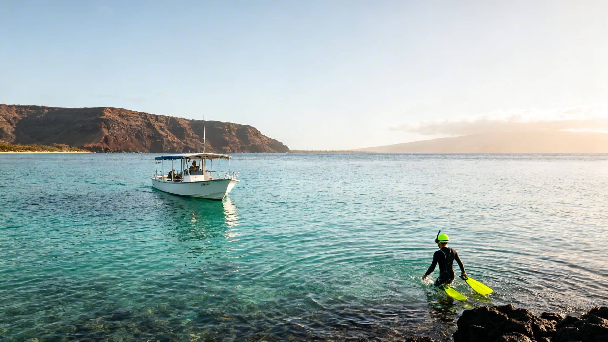 A snorkeler with yellow fins enters clear blue water from a rocky shore, with a boat and mountainous coastline.
