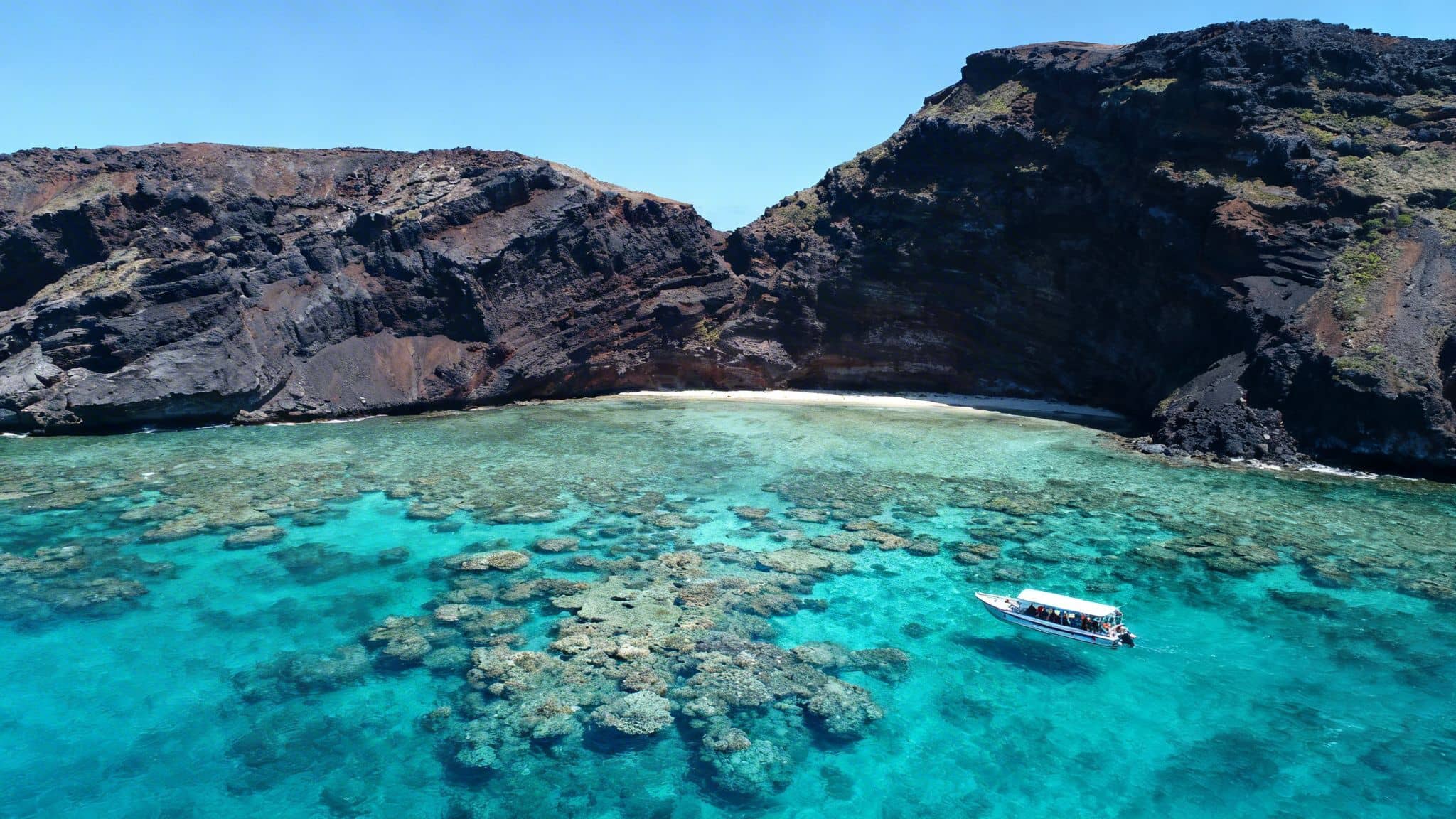 Aerial view of a tour boat on vibrant turquoise water with coral reefs near dark volcanic cliffs and a sandy beach.