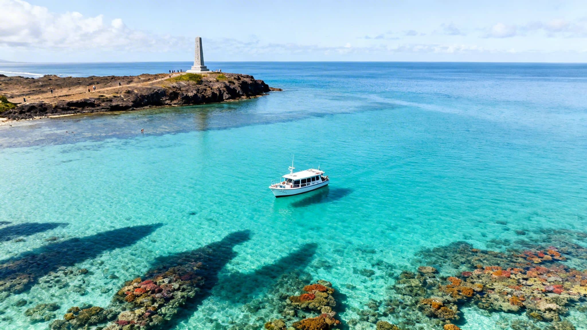 Aerial view of a white boat in turquoise water with colorful coral reefs, a monument, and people on a rocky coast.